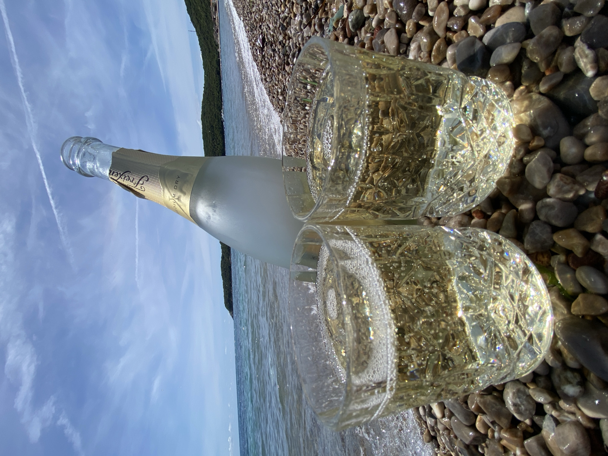 The image shows a bottle of sparkling wine and two glasses filled with it, positioned on a pebble beach, likely in Prijevor, Montenegro. The bottle is laying horizontally, slightly tilted, with the neck pointing towards the sky. The glasses are near the bottle, nestled among the pebbles. The sea water laps at the edge of the pebbles. In the background, there is a shoreline and a blue sky with some clouds.