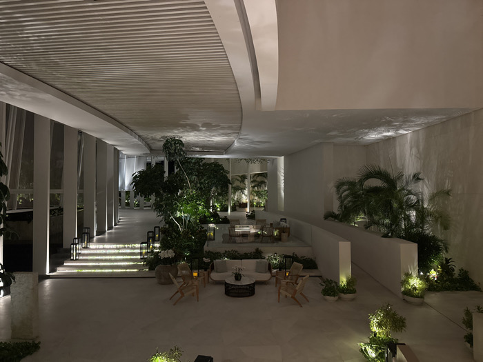 A spacious, modern outdoor lounge area features white tiled flooring and ample natural light. Lush greenery, including trees and palms, complements the minimalistic white architecture. Seating arrangements consist of sofas and chairs around coffee tables. Steps with lighting lead to a raised platform with additional seating. The ceiling has a partially curved, slatted design. The scene evokes a serene and luxurious ambiance.