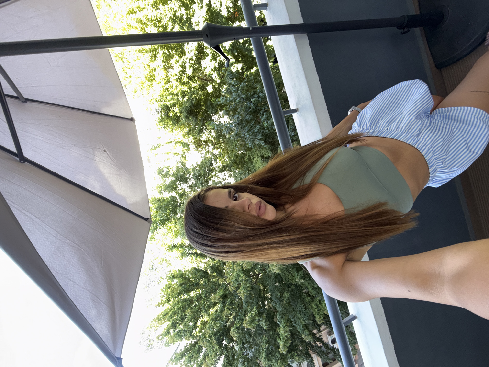 In a selfie taken on a balcony in Plovdiv, Bulgaria, a woman with long brown hair is posing. She is wearing a green sports bra and light blue and white striped shorts. Behind her, lush green trees and a portion of a white building are visible. A large patio umbrella, partially open, stands to her right. The railing of the balcony is a dark grey metal, and the floor appears to be carpeted. The overall scene suggests an outdoor setting, possibly a residential balcony, on a bright day.