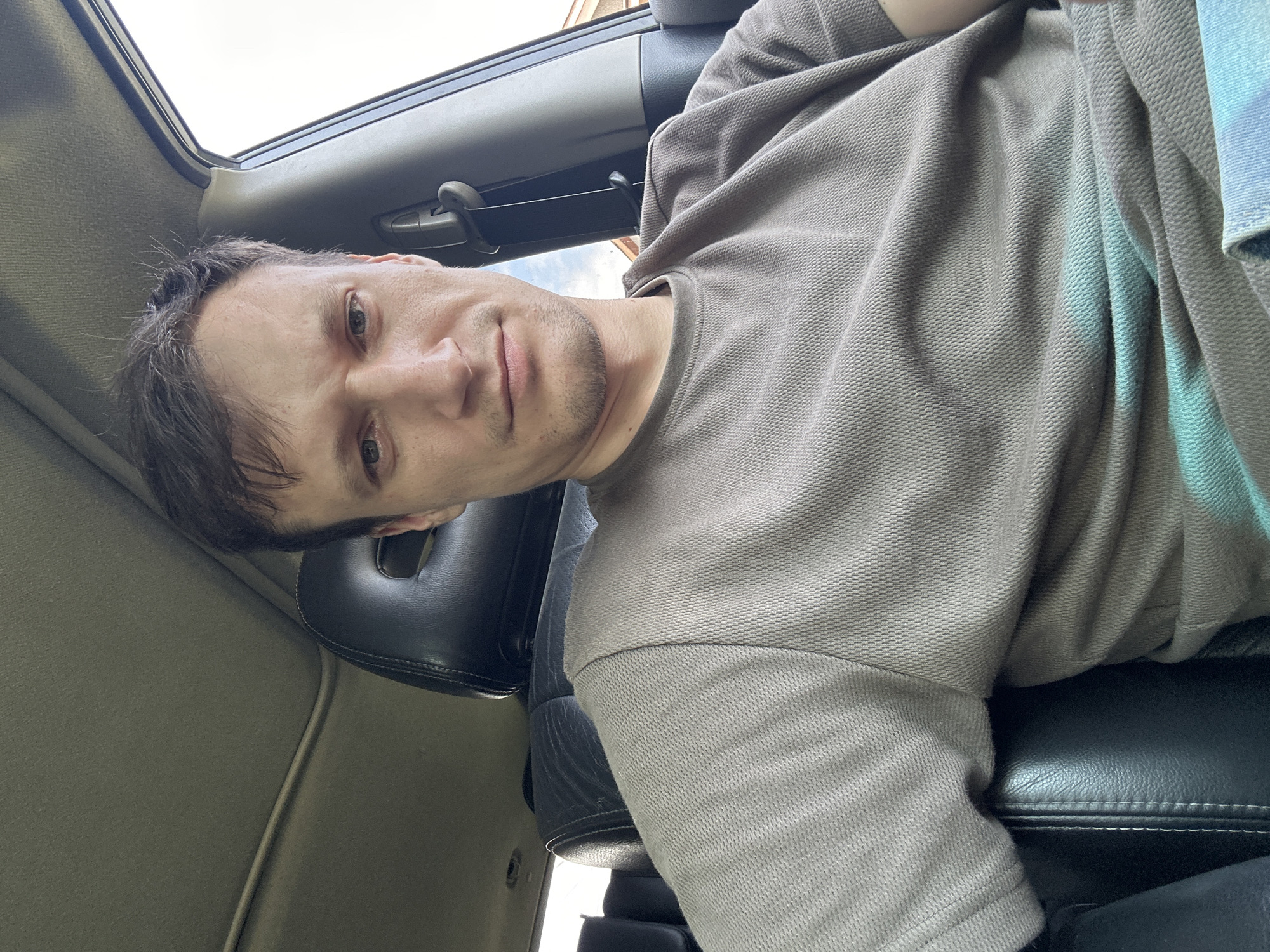 A man with short, dark brown hair and light skin is seated in the driver's seat of a car. He is wearing a short-sleeved, brown, textured t-shirt. His head is turned towards the camera, and he is looking directly at it with a neutral expression. The interior of the car is visible, including the headrest of his seat and part of the car's ceiling. The background shows a cloudy sky and some houses, suggesting an outdoor setting within the city of Makiv, Ukraine. The image appears to be a selfie taken from the man's perspective.