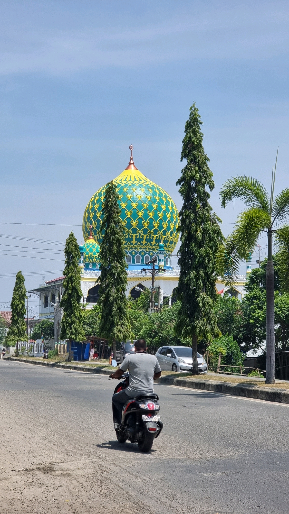 A medium shot shows a person riding a red and black scooter from behind on a paved road in the city of Lhokseumawe, Indonesia. The rider is wearing a gray t-shirt and dark pants. The scooter's license plate is partially visible. In the background, a large mosque with a prominent green and yellow dome is visible. The mosque features ornate detailing on its dome and lower structures. Several tall, slender green trees are positioned in front of the mosque, along with other foliage. A silver car is parked on the side of the road. The sky is clear and blue, indicating a sunny day. Overhead power lines are also present.