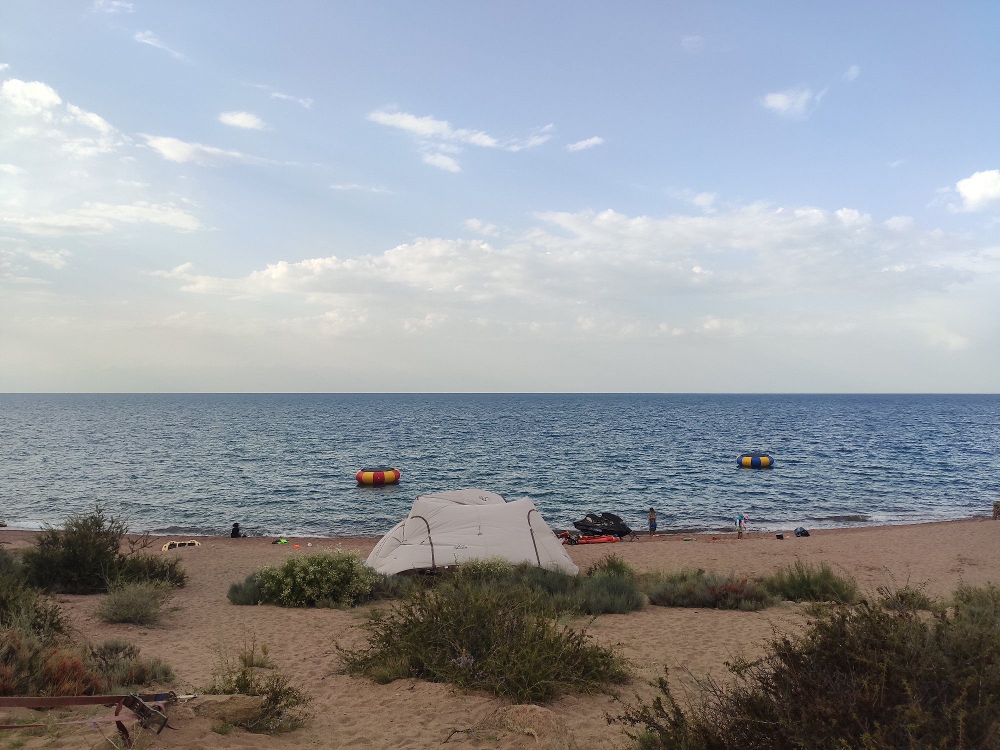 The image depicts a sandy beach with a body of water stretching to the horizon under a partly cloudy sky. In the foreground, several bushes and sparse vegetation are scattered across the sand. A light-colored tent is set up on the beach, with a jet ski and a red inflatable raft nearby. Further down the beach, a few individuals are visible, some near the water and others on the sand. In the water, there are two inflatable objects, one a yellow and blue ring and the other a yellow and red ring. The scene suggests a recreational setting by a lake or sea.