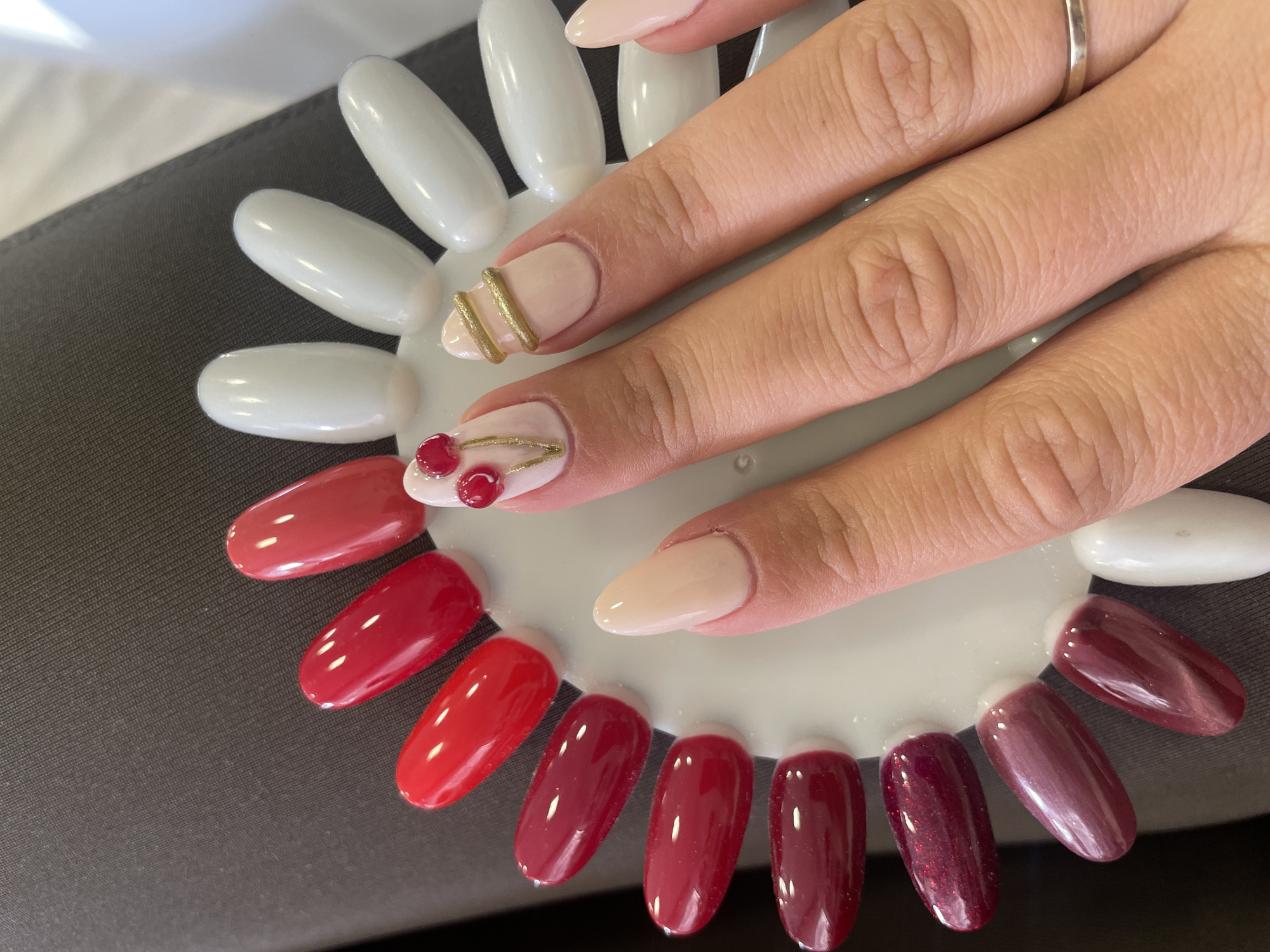A close-up shot features a hand resting on a palette showcasing various nail polish colors, arranged in a semi-circle. The hand's fingernails are painted in a nude shade, except for the ring finger and middle finger, which have nail art. The ring finger has a design incorporating small red cherry-like adornments connected by gold chains, and the middle finger features two parallel gold bars. The palette has shades ranging from light red to deep burgundy. The palette itself is placed on a dark gray surface.
The location is unknown.