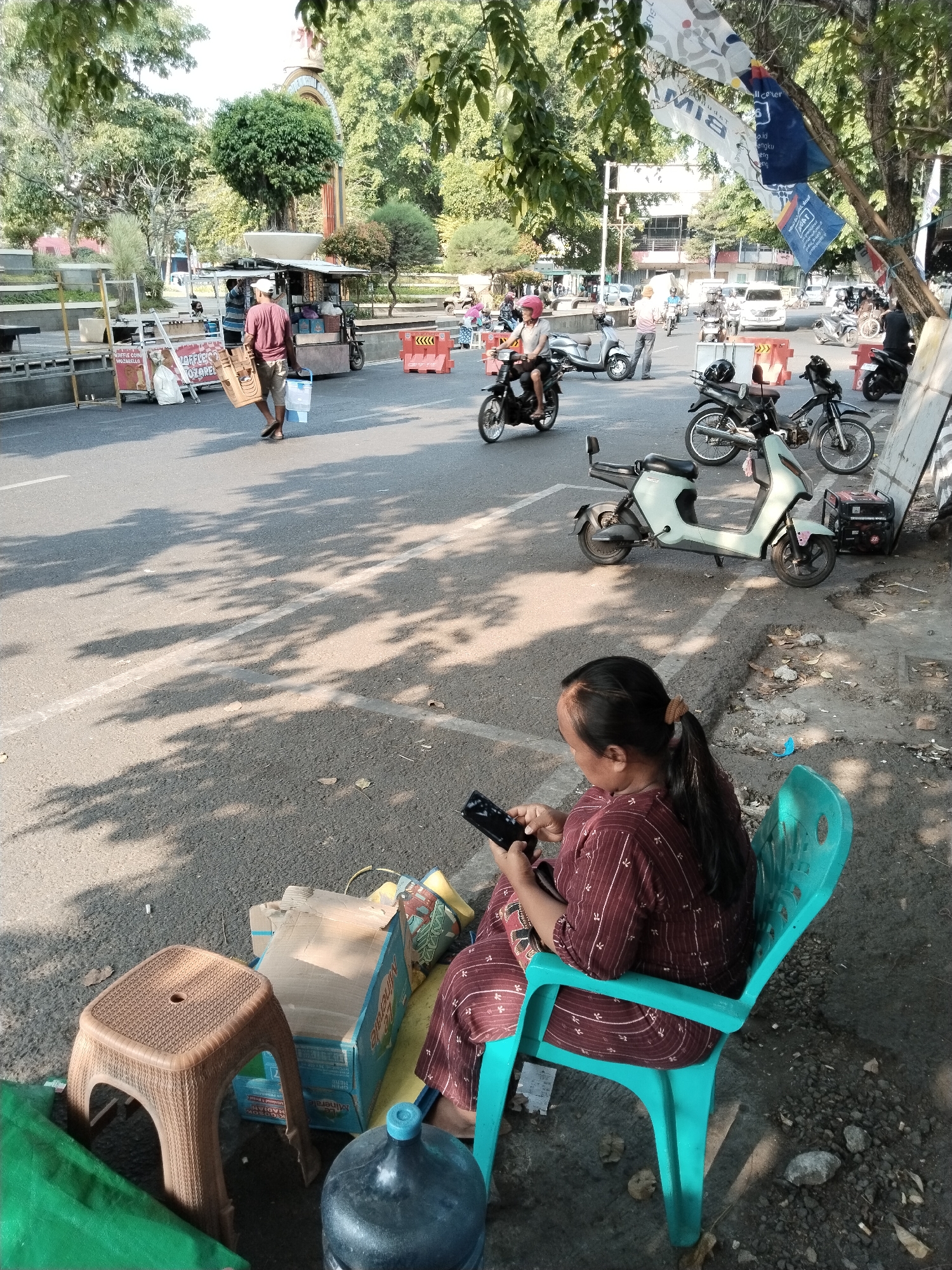 The image shows a street scene in Kecamatan Pati, Indonesia, on a sunny day. In the foreground, a woman with her hair in a ponytail is sitting on a teal plastic chair and looking at her phone. She is wearing a maroon and white patterned outfit. Next to her is a small brown plastic stool and a large blue water jug. Behind the woman, there are boxes and a green tarp. In the middle ground, several motorcycles and scooters are parked on the side of the road. One scooter is light green, and another is black. Further back, a man is walking across the street carrying a crate and a blue container. He is wearing a maroon shirt and shorts. There are food stalls and other vendors lining the side of the road. In the background, trees and buildings are visible, and there are some flags hanging from a tree. The street is paved and has white lane markings. The overall atmosphere is that of a busy urban street with economic activity.