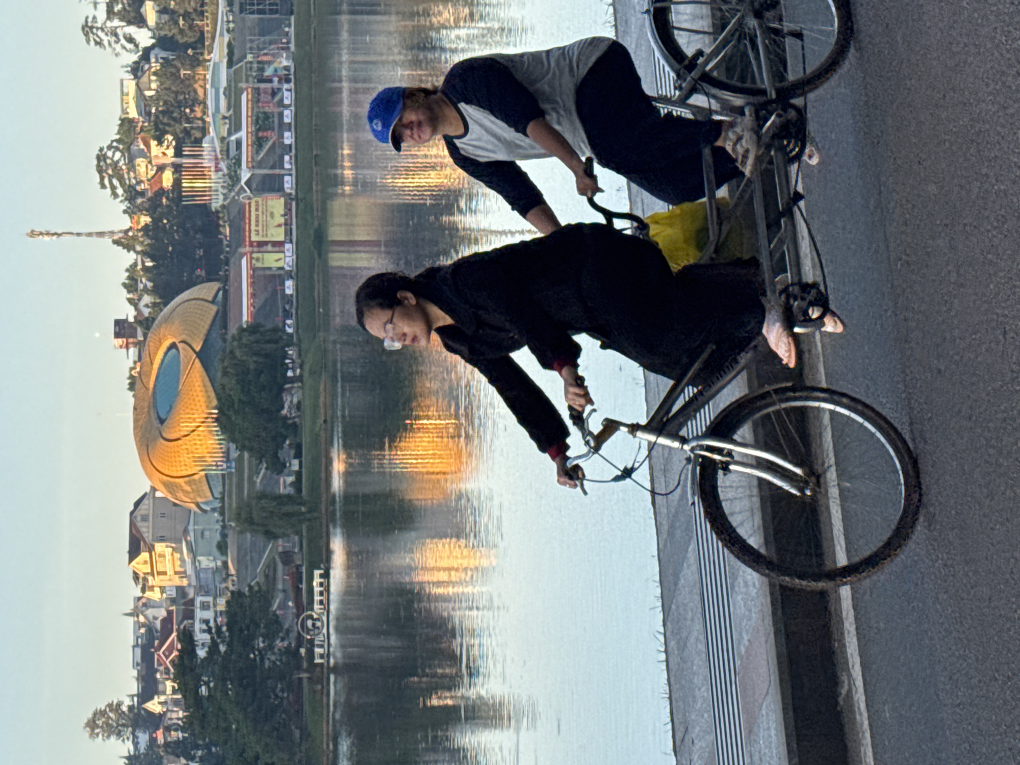 Two individuals are riding a tandem bicycle along a paved path next to a body of water, with a city skyline in the background. The person in front, wearing glasses and a black jacket, is pedaling. The person behind, wearing a blue baseball cap and a grey and black shirt, is also pedaling. A yellow bag is visible on the rear of the bicycle. The water reflects the buildings and trees of the city, which appears to be Phường Xuân Hương - Đà Lạt, Vietnam, based on the distinctive architecture of a dome-shaped building in the distance. The sky is clear and appears to be sunrise or sunset, casting warm light on the scene.