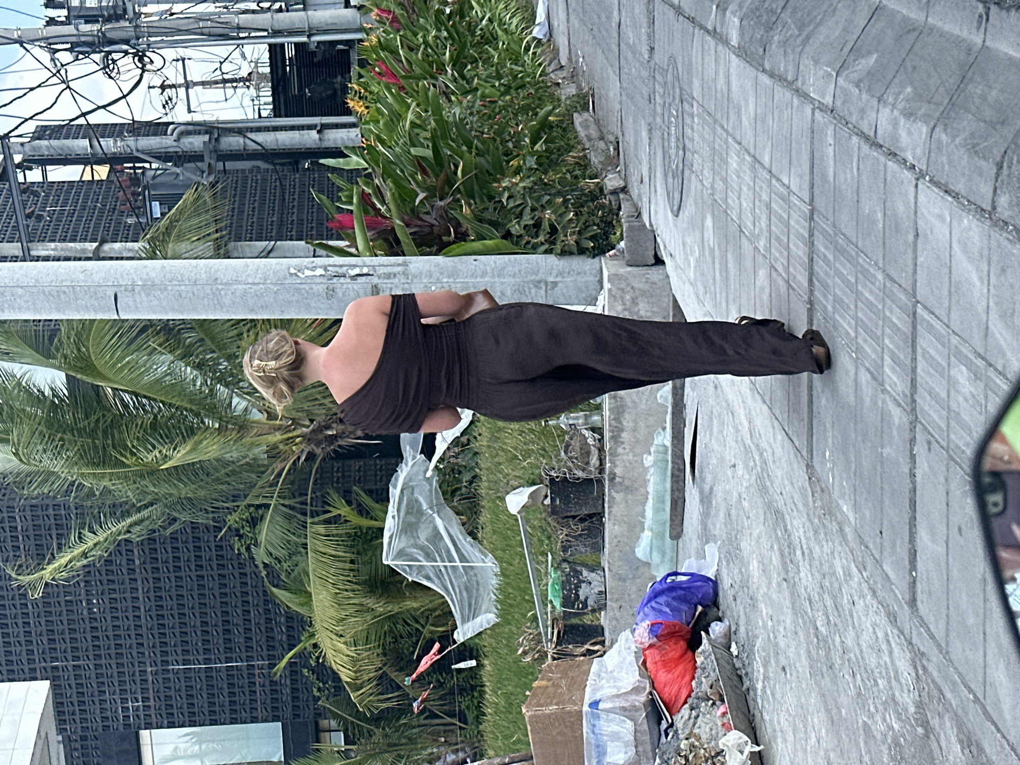 The image shows a woman from behind, walking on a sidewalk in North Kuta, Indonesia. She is wearing a dark brown one-shoulder top and matching dark brown pants. Her blonde hair is pulled back in a bun. The background consists of a modern building with a dark patterned facade, palm trees, lush green foliage with pink and red flowers, and utility poles with numerous wires. The sidewalk is paved with grey tiles, and there is a concrete curb and gutter. To the left of the woman, on the sidewalk, is a pile of debris including trash bags, plastic sheeting, and what appears to be construction materials. The scene suggests an urban environment that may be experiencing some littering or ongoing construction.