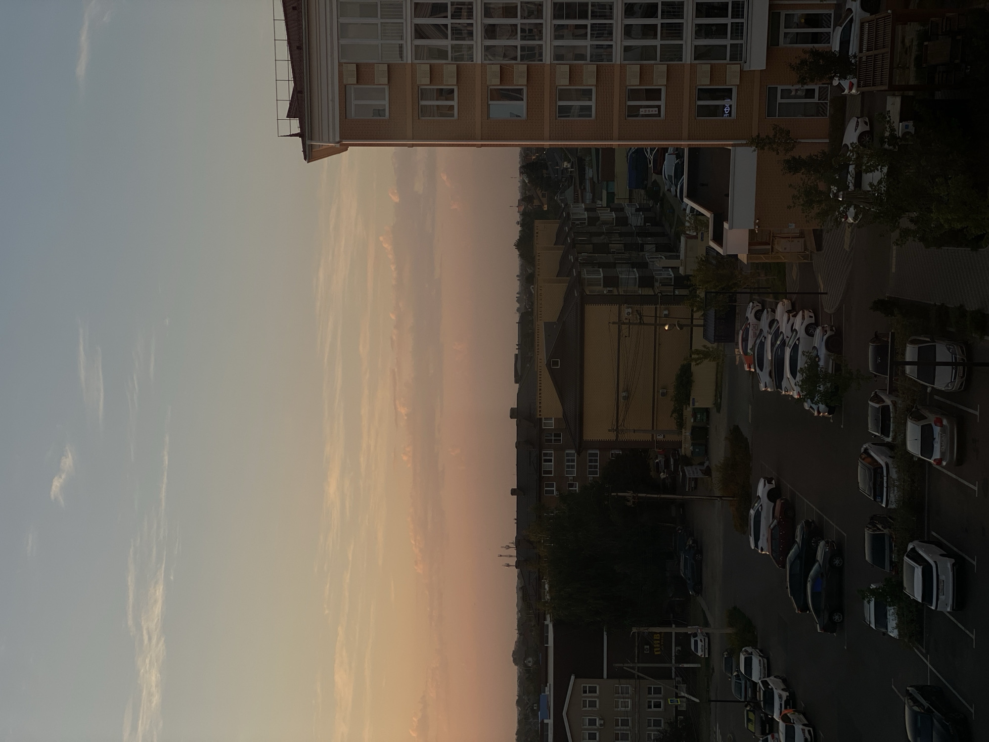 The image is an exterior, elevated, high-angle shot of a residential area in Zelenopol'skii, Russia, during twilight. The sky is a gradient of pale blue at the top, transitioning to a soft peach and pink hue near the horizon, with wispy clouds scattered across it. In the foreground and mid-ground, various buildings are visible, including a prominent multi-story brick apartment building on the right with numerous windows and balconies. Several other apartment buildings and houses, varying in height and architectural style, are situated throughout the scene. A parking lot is visible in the lower half of the image, containing numerous parked cars of various makes and models. Trees and foliage are interspersed among the buildings and parking areas. The lighting suggests it is either sunrise or sunset, casting long shadows and a warm glow over the landscape. There are no visible people or active interactions depicted in the scene.