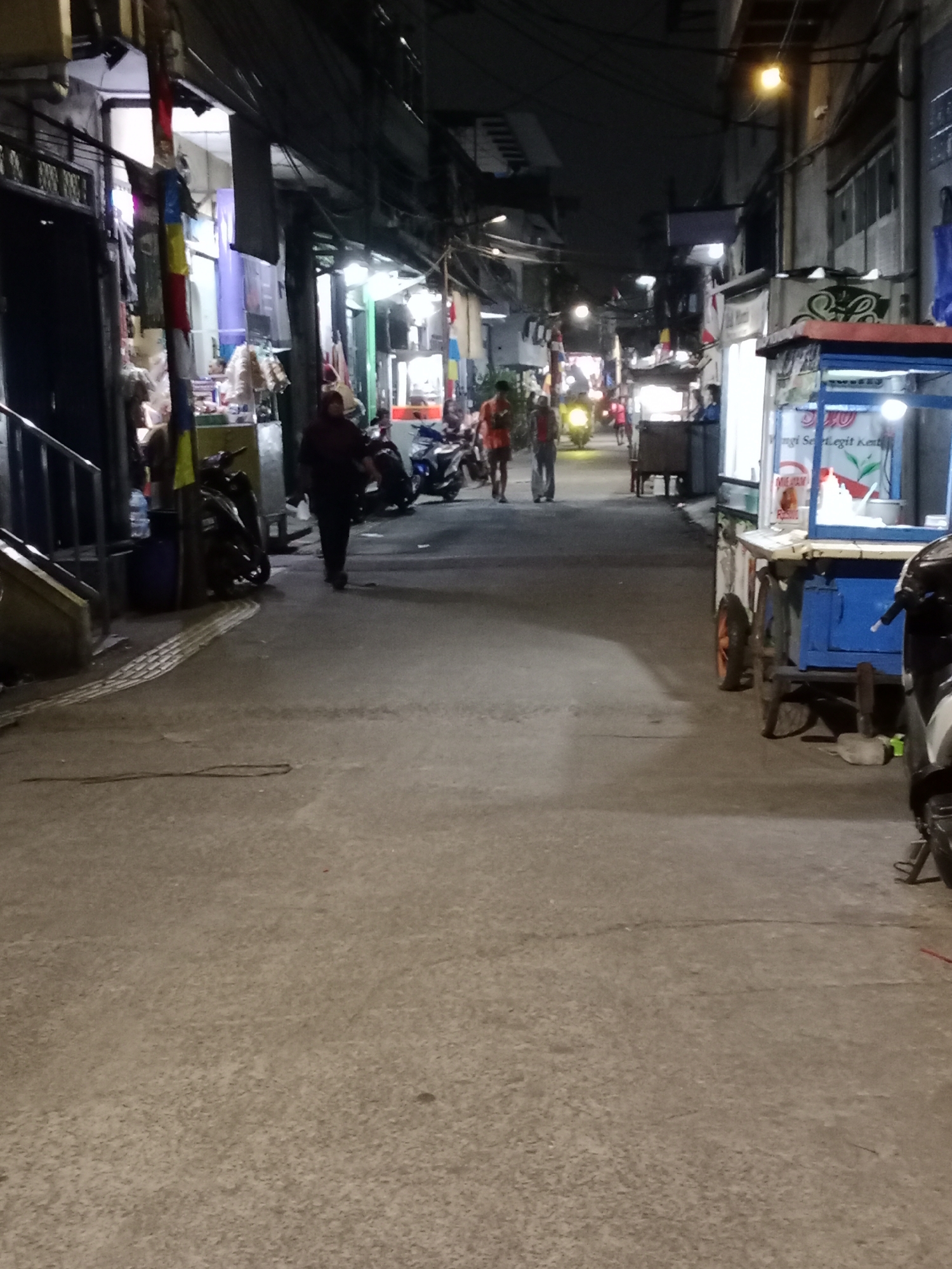 The image depicts a street scene at night in West Jakarta, Indonesia. A paved road runs through the middle of the frame, with buildings lining both sides. Several motorcycles are parked along the left side of the street. A person wearing dark clothing walks away from the camera on the left side of the road. Further down the street, two individuals are walking towards the camera. One is wearing a red shirt and the other is in a light-colored shirt. A small food cart with a blue frame and a white awning is visible on the right side of the road, illuminated by lights. The environment appears to be a typical urban street with overhead wires and various shopfronts.
