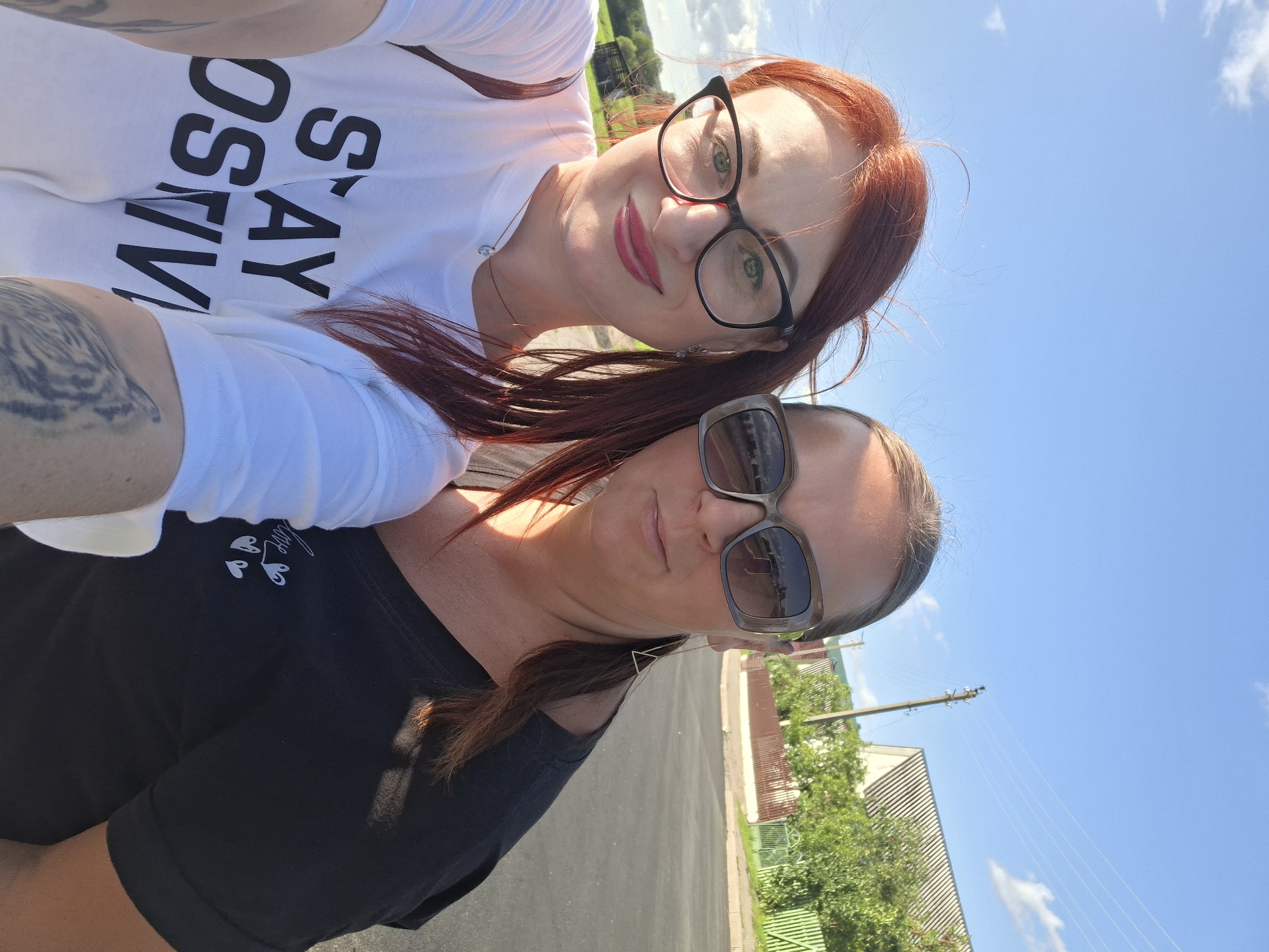 The image is a selfie taken outdoors, with two women in the foreground. The woman in the front is wearing a black t-shirt and sunglasses, with her hair pulled back in a ponytail. She is looking directly at the camera with a slight smile. The woman behind her is wearing a white t-shirt with black text that says "STAY MASO." She has red hair, glasses, and is also smiling at the camera.
The background shows a blue sky with some white clouds, suggesting it is daytime and the weather is clear. There are trees and buildings visible, indicating an outdoor setting, possibly a residential area. There are also power lines visible, consistent with a rural or suburban location. The overall mood of the image appears to be happy and relaxed, with both women smiling. The image is taken in Myadzyel, Belarus.