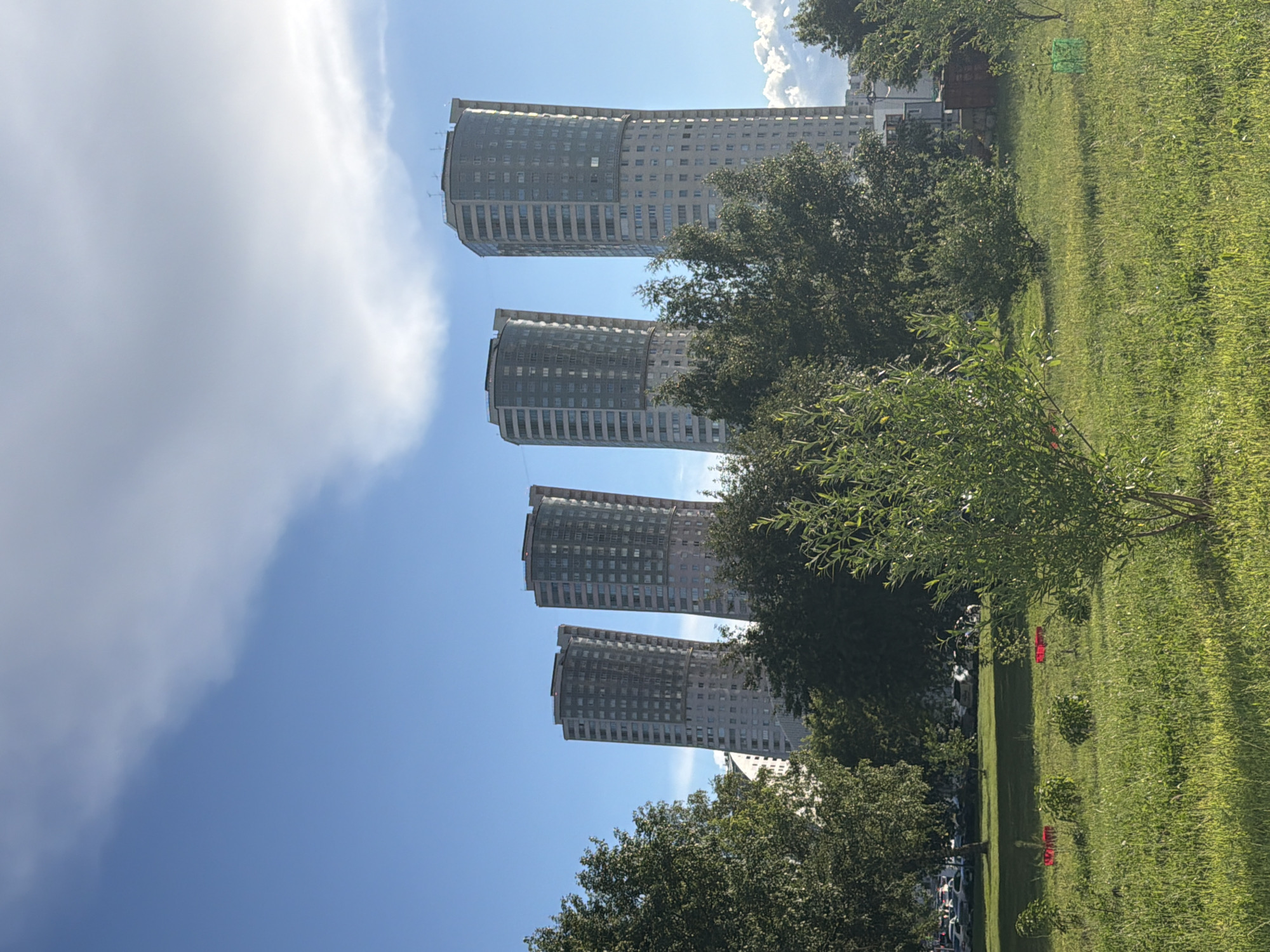The image shows a row of tall, modern buildings with curved facades, set against a bright blue sky with wispy white clouds. The buildings are a uniform light gray color and are lined with many windows. Lush green trees and manicured grass surround the buildings, creating a park-like setting. The sun appears to be shining, casting shadows across the grass and illuminating the scene, suggesting it is daytime. There are no people visible in the image. The scene appears to be an urban environment with residential or office buildings integrated with green spaces. The overall impression is one of a bright, sunny day in a well-maintained urban area.