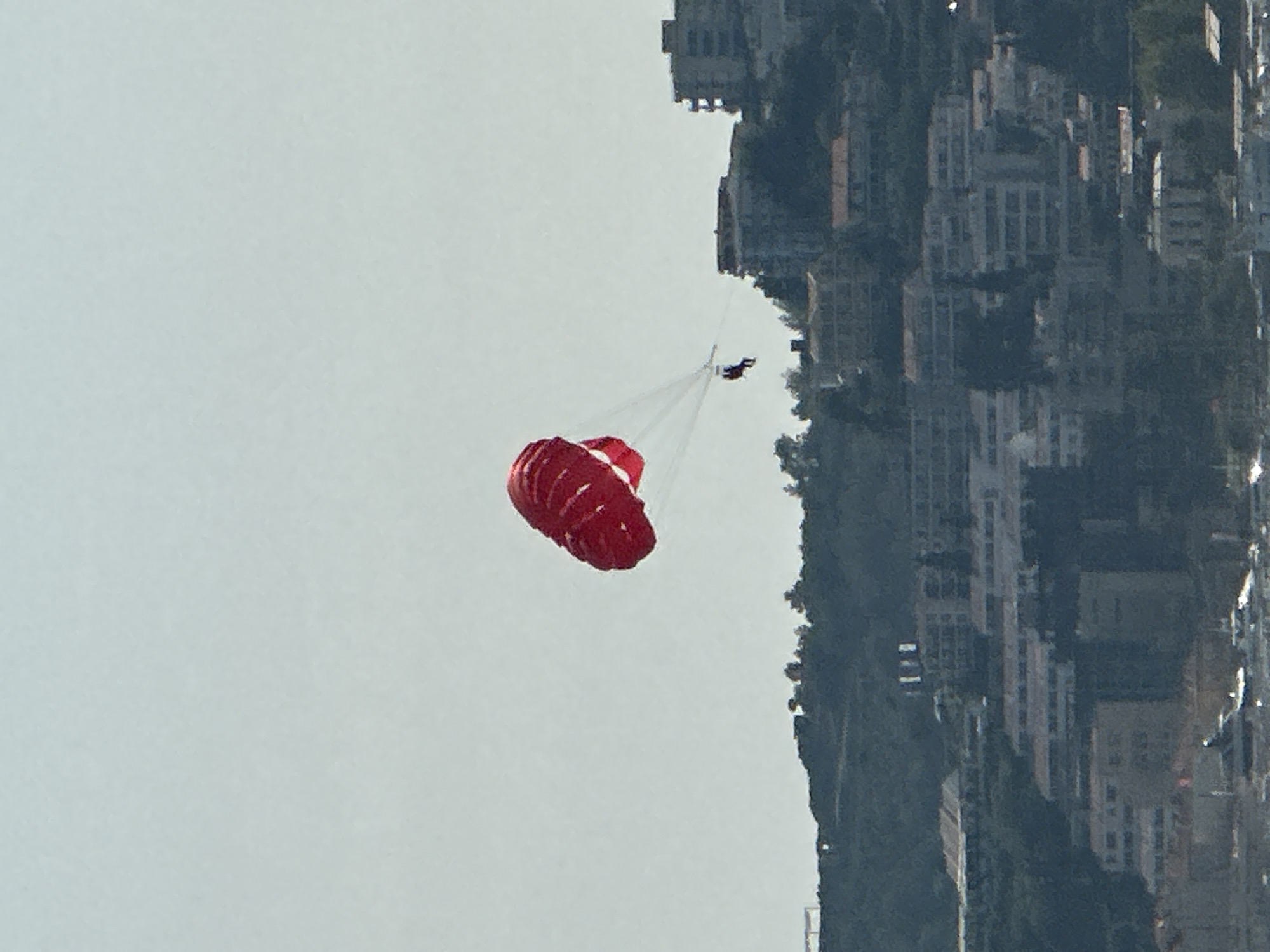 An aerial view captures a person parasailing with a red parachute against a hazy sky. Below the parachute, a densely populated coastal town is visible, built on a hillside overlooking the Adriatic Sea. The town features numerous buildings with white and light-colored facades, interspersed with lush green trees. In the foreground, a body of water with several boats and what appears to be a seaplane suggests a harbor or marina. The overall scene depicts a sunny day with limited visibility due to atmospheric haze. The town's architecture and terraced layout strongly suggest it is Budva, Montenegro, a popular tourist destination known for its beaches and old town.