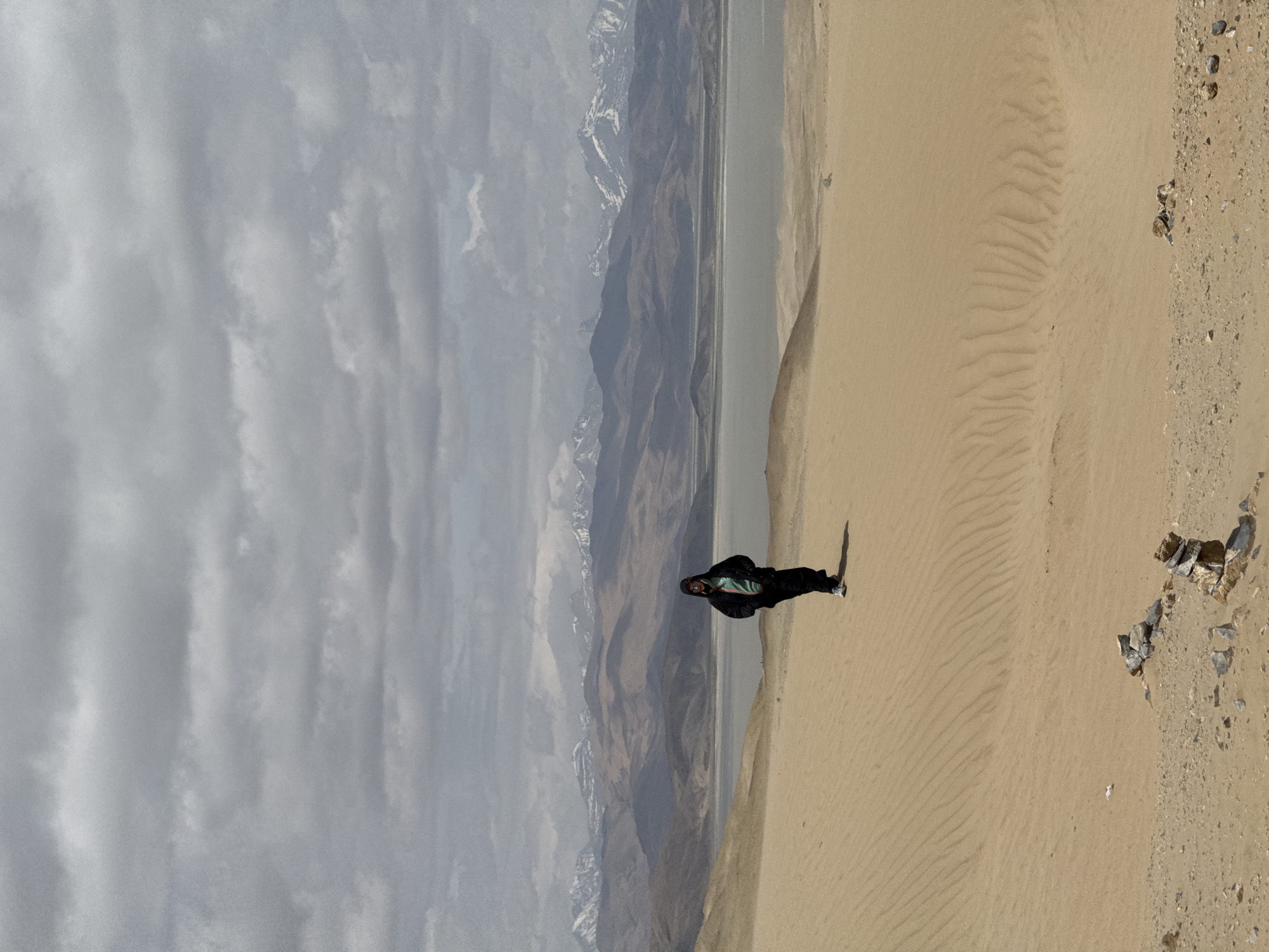A person stands on a sand dune. Behind the sand dune are mountains, clouds, and a body of water. The scene appears to be in a remote location. The photo may have been taken in or near Labrang township in China.