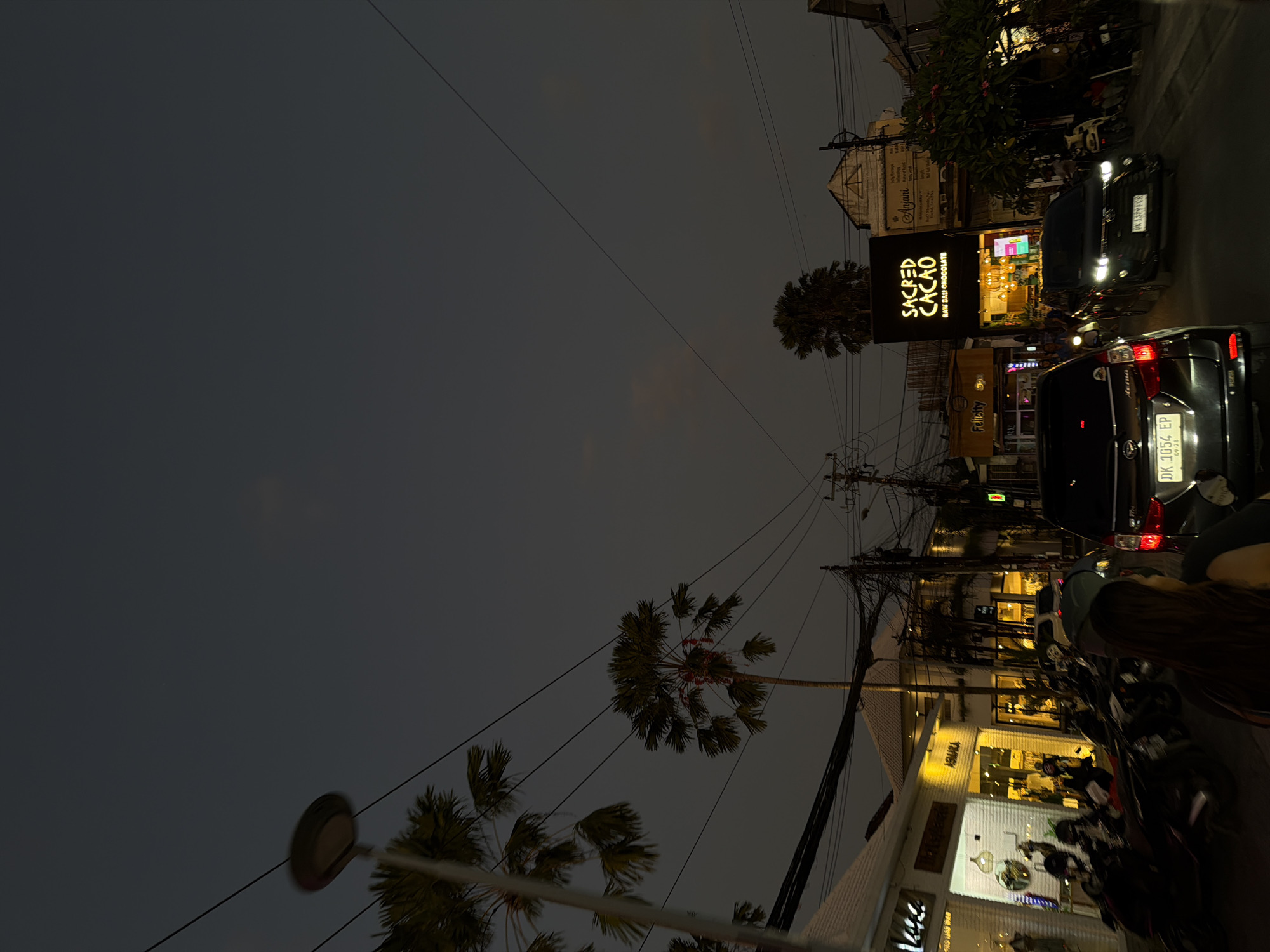 This image depicts a street scene at dusk or night in North Kuta, Bali, Indonesia. The sky is a dark grey, indicating the absence of daylight. The street, which appears to have at least two lanes, is illuminated primarily by artificial light sources from shops and vehicles.
In the immediate foreground, the back of a person's head and right shoulder, featuring long dark hair, is visible on the left side. Directly in front of the viewer, a dark-colored SUV with Indonesian license plate `DK 1054 EP` is parked or stationary. Its red taillights are visible.
Along the left side of the street, a series of commercial establishments are brightly lit, showcasing their interiors and merchandise. Visible signs include "ADARA" and partially visible "Kees". Numerous motorcycles are parked along the sidewalk in front of these shops.
On the right side of the street, another dark-colored SUV, with license plate `DK 3329 KCK`, is moving away from the viewer, its headlights illuminated. Behind it, a prominent black storefront sign reads "SACRED CACAO RAW BALI CHOCOLATE" in white lettering, with bright yellow lights illuminating its entrance. Adjacent to Sacred Cacao, another building has a sign, partially obscured, that appears to be "Anjani". Another distant sign reads "Felicity".
Multiple tall palm trees are silhouetted against the dark sky. One palm tree in the center has red festive decorations wrapped around its trunk. Numerous overhead utility wires form a dense network across the upper midground and background, connecting to visible utility poles. A blurred street light is visible above a palm tree on the far left. The architectural style of the buildings includes tiled roofs and brightly painted facades for the commercial premises.