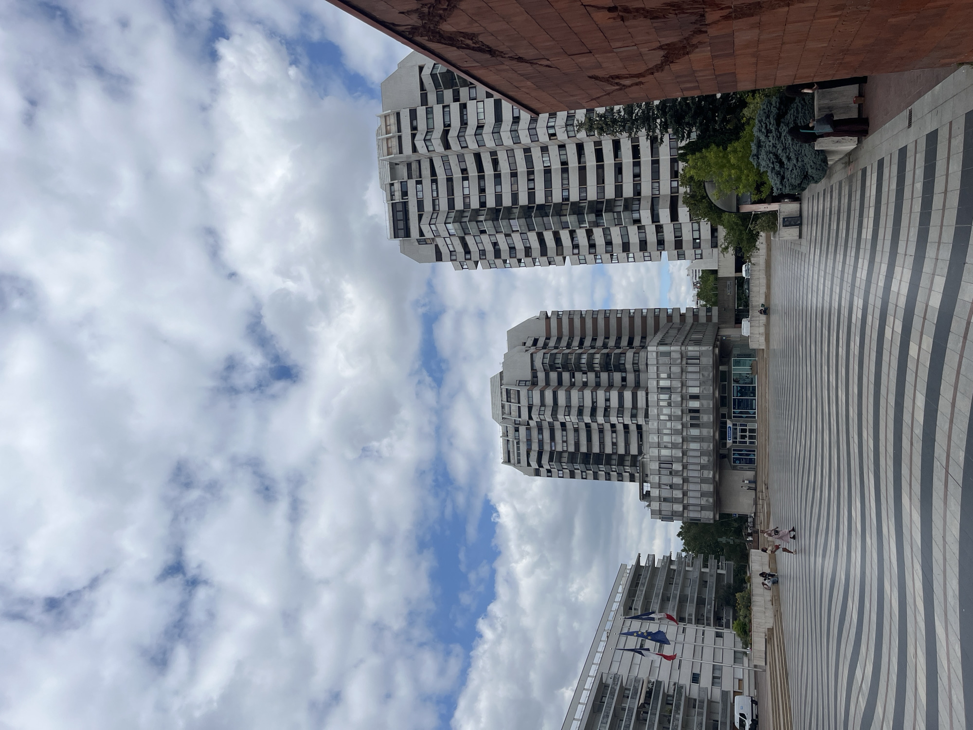 This media file shows an outdoor scene in Créteil, France, featuring modern architecture and a cloudy sky. In the foreground, a patterned pavement with alternating dark and light stripes leads towards two tall, white, multi-story buildings with numerous windows and balconies. The buildings have a distinctive stepped or curved facade.
In the middle ground, several people are visible. Some are sitting on a stepped area in front of the buildings, while others are standing and walking on the patterned pavement. There are also some trees and greenery interspersed with the buildings. A large, reddish-brown textured surface, possibly part of another building, is visible in the upper left corner, contrasting with the smooth facades of the residential towers.
In the background, more buildings and trees are visible, suggesting an urban environment. Several flags, including what appears to be the European Union flag and the French flag, are displayed on poles in front of one of the buildings. A white van is parked near the flags.
The sky is filled with a mixture of white and grey clouds, with patches of blue sky peeking through, indicating a partly cloudy day. The lighting suggests it is daytime, likely late morning or afternoon. The overall mood of the scene appears relaxed and casual, with people enjoying the outdoor space. No discernible text is visible in the image.