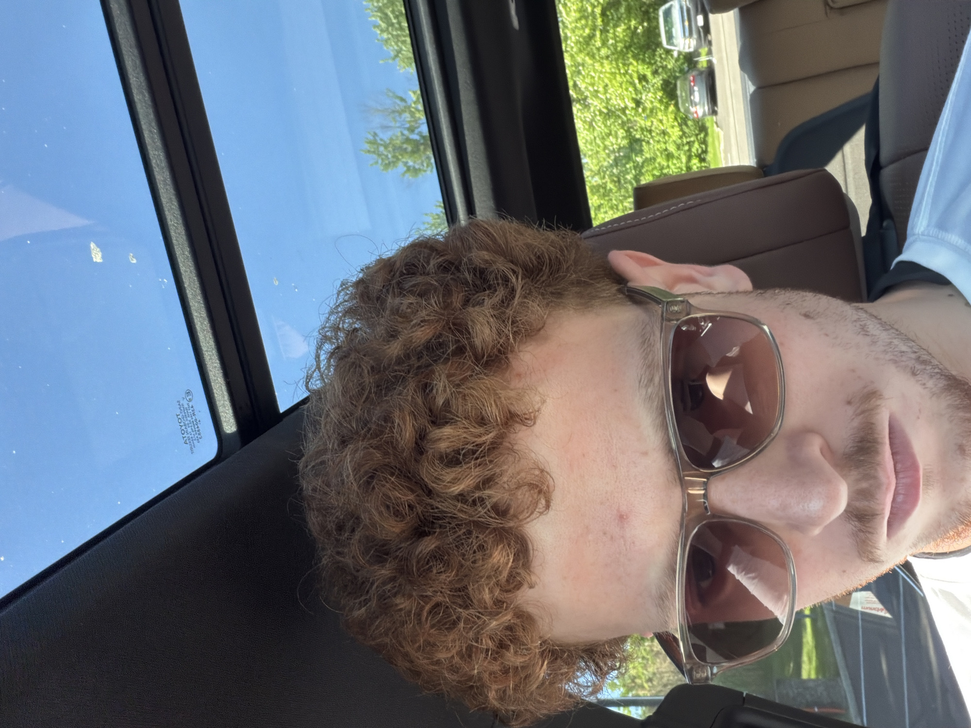 A young person, likely male, with prominent curly, reddish-brown hair and fair skin, is depicted in a selfie-style shot from inside a car. The person is wearing sunglasses with clear frames and brown lenses, which obscure their eyes but reflect the car's interior. A slight mustache and chin hair are visible. They are wearing a light-colored, possibly white or light blue, collared shirt.
The setting is inside a car, indicated by brown leather seats and black interior trim visible around the person. The sun is shining brightly, suggesting daytime. A panoramic sunroof above the person reveals a clear blue sky and distant green trees, along with some visible dirt or spots on the glass. Through the side windows, more green foliage and reflections of other vehicles on a paved surface can be seen, hinting at an outdoor environment, possibly a parking lot or road.
Notable details include reflections of the car's interior and potentially the camera device in the sunglasses lenses. Inverted text, "ΔTOYOT" (likely a backward "TOYOTA" with a delta symbol), and other unreadable manufacturing marks are visible on the sunroof glass. The overall impression is a casual moment captured in good weather. The location context is Beachwood, United States of America.