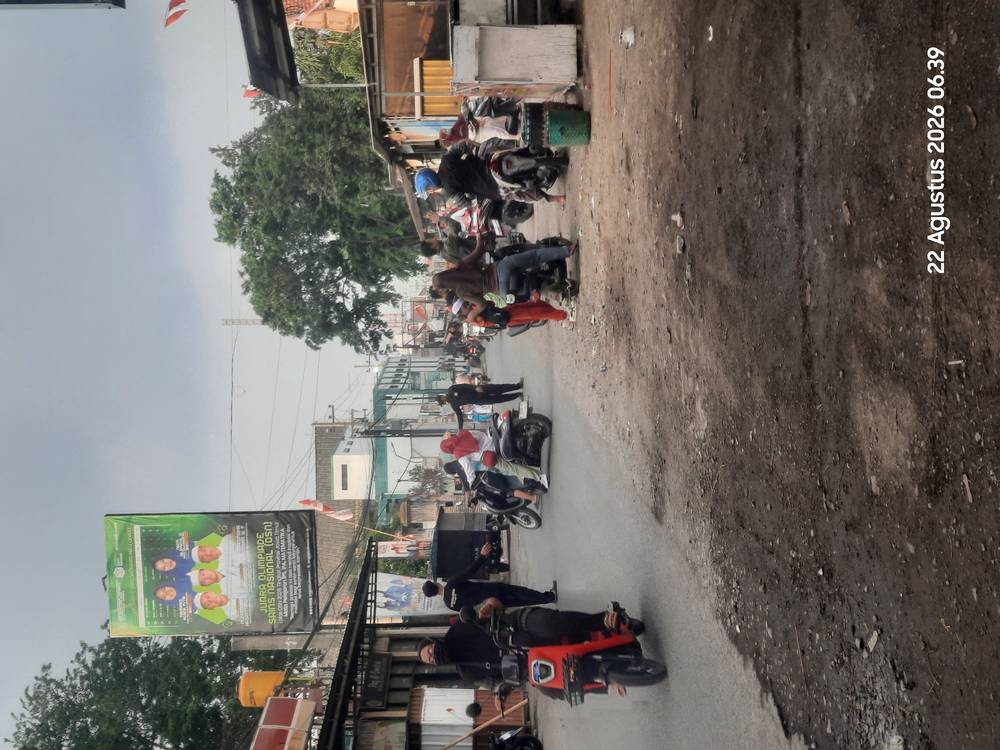 The image depicts a street scene in Pajarakan Kulon, Indonesia. The foreground shows a dirt and asphalt road with debris. In the middle ground, several people are on or near motorcycles. On the left, a woman wearing a black hijab and mask is riding a red electric scooter. Further down the street, a group of individuals are gathered around motorcycles. A large banner hangs on a pole to the left, displaying images of people and text in Indonesian. The background features buildings, trees, and a cloudy sky. A timestamp "22 Agustus 2026 06.39" is visible in the bottom right corner.