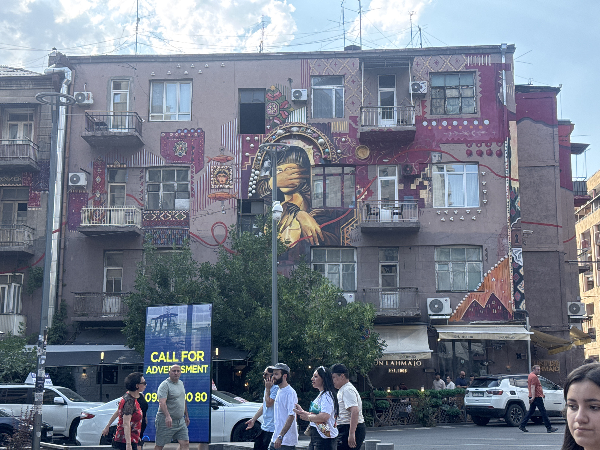 The image depicts a street scene in Yerevan, Armenia, focusing on a building adorned with a large, vibrant mural. The mural covers a significant portion of the building's facade, featuring abstract patterns, geometric designs, and figurative elements, including a woman's face partially obscured by her hand. The building itself appears to be an apartment complex with balconies and windows, some of which are air-conditioned.
In the foreground, several people are visible walking on the sidewalk. To the left, a tall, blue digital billboard displays the text "CALL FOR ADVERTISEMENT" in yellow, along with a phone number. A white car and a black SUV are parked on the street. The street is paved, and there are trees and awnings of businesses along the storefronts on the ground floor of the building. The sky is partly cloudy. The overall impression is a lively urban environment with a strong artistic element integrated into the architecture.