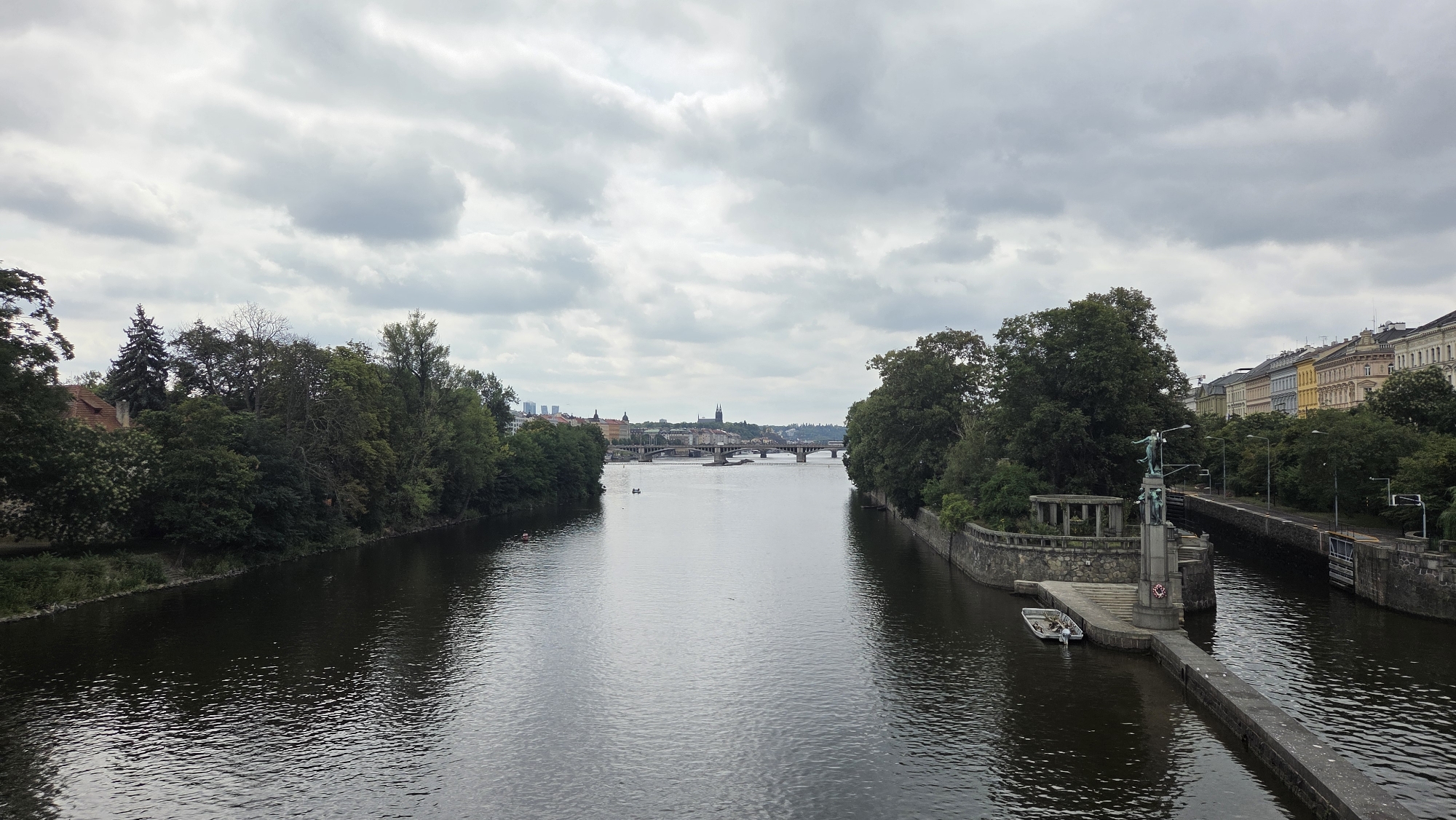 The image shows a wide river flowing through a city with a cloudy sky overhead. On the left bank, lush green trees and a building with a red tiled roof are visible. The river widens as it approaches a bridge in the distance, which spans across to the right bank. The right bank features a stone embankment with a monument topped by a statue of a winged figure, accompanied by smaller statues. A small boat is docked near the monument. Further along the right bank, a series of colorful buildings with multiple stories are visible, interspersed with trees. The scene is likely Prague, Czechia, given the architectural style and the presence of a significant river.