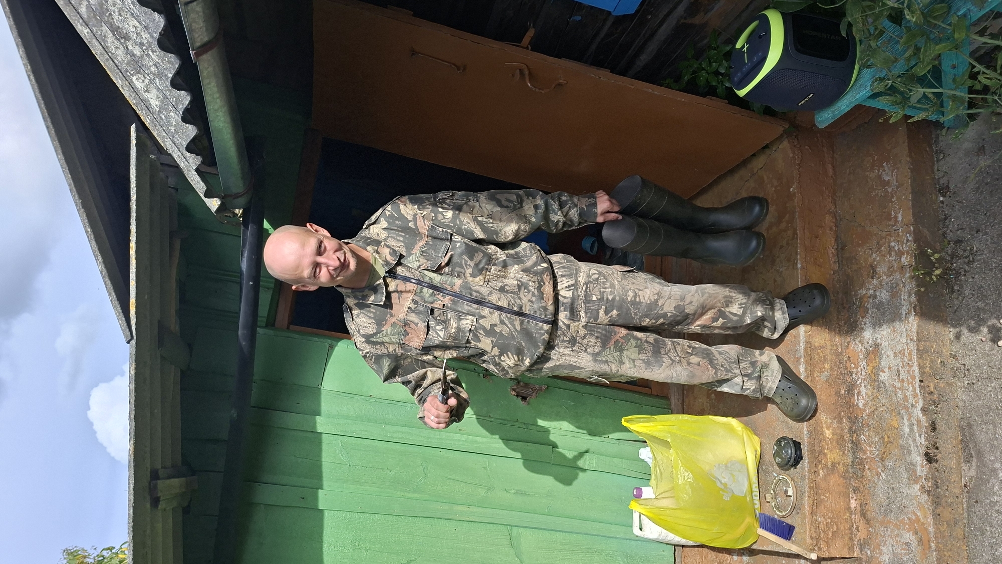 A bald man wearing camouflage clothing, dark crocs, and a green t-shirt stands on the porch of a rural wooden building with a green exterior. He holds a knife in his right hand and a pair of black rubber boots in his left. In front of him on the porch are a yellow plastic bag, a blue brush, a round metal object, and a black cap. Behind him, a dark brown door is ajar, revealing an interior. To the right of the door, on a blue stool, sits a black and neon green portable speaker. The building appears to be in disrepair, with peeling green paint on the walls and a dilapidated roof. The porch steps are made of rough, weathered wood. The ground is dirt and scattered debris. The sky above is partially cloudy. The location is likely in the Svatkaŭski Sel'ski Savec, Belarus, suggested by the rustic rural setting.