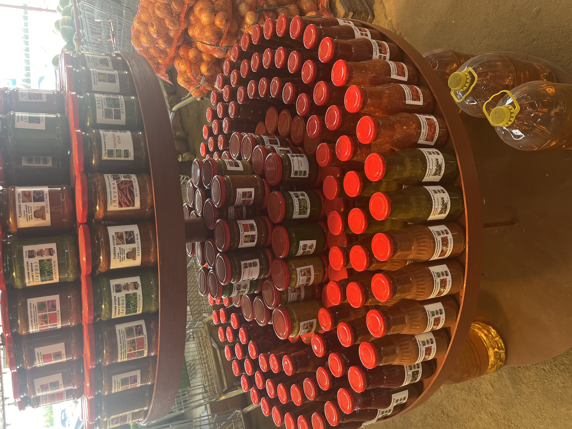 The image depicts a retail display of assorted preserved goods, primarily in glass jars and plastic bottles, arranged on tiered circular shelves. The products, predominantly appearing to be sauces, jams, or pickled vegetables, feature red lids and labels with Cyrillic text. Some labels display images of ingredients and contact information. Prominently displayed in the background are two large transparent plastic containers filled with a golden liquid, possibly oil or juice, with yellow lids and handles. To the right of the shelving unit, several netted bags filled with onions are stacked. The setting appears to be an outdoor or open-air market stall, with a concrete or paved ground surface. A metal shopping cart is partially visible in the upper left corner. The overall lighting suggests an overcast day. Based on the provided context, this scene is located in the city of Plastunovskaya, Russia. There are no people visible in the frame.