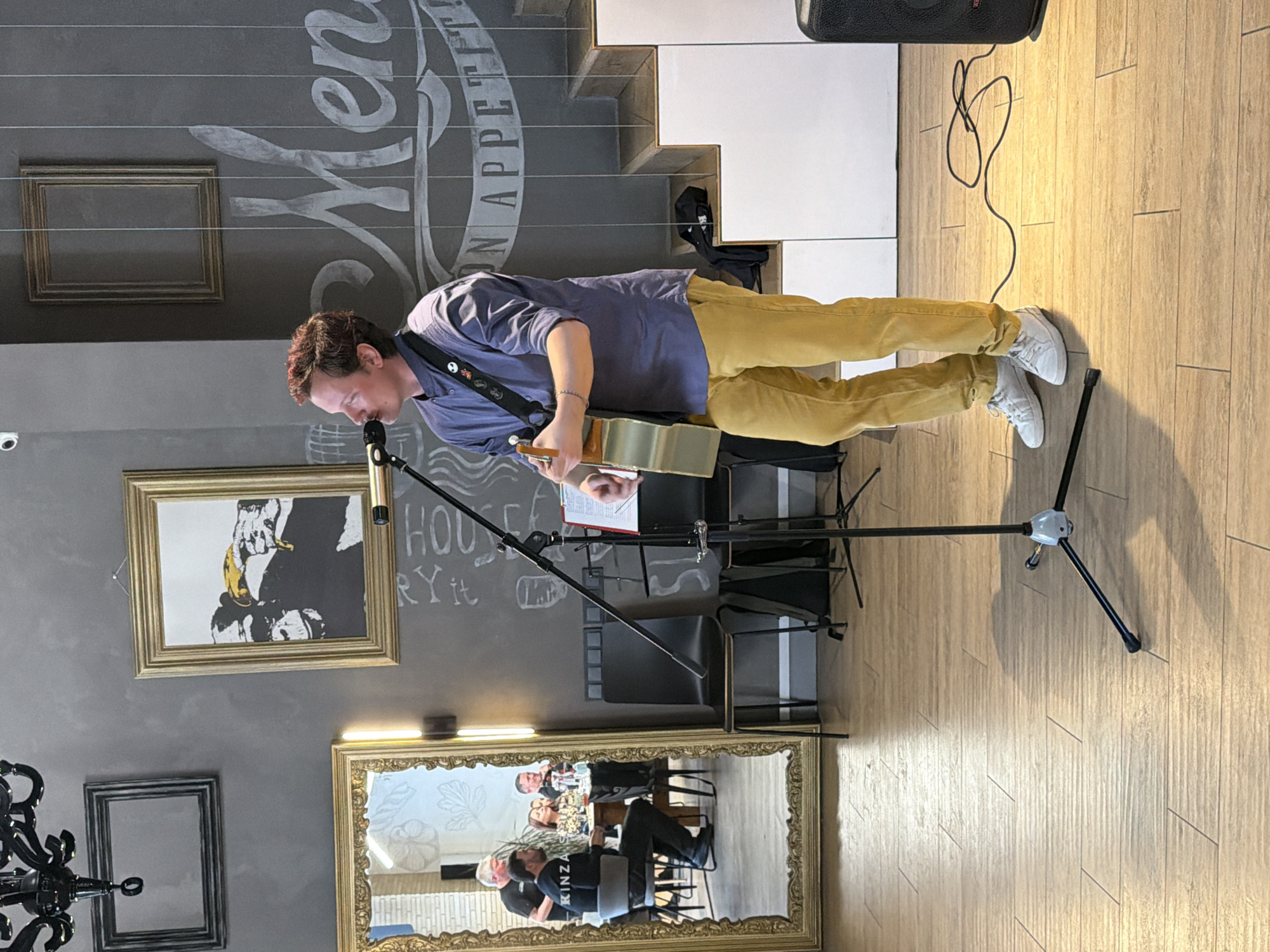 A man is standing and playing an acoustic guitar into a microphone in a room that appears to be a cafe or restaurant. The man is wearing a purple shirt, yellow pants, and white sneakers. Behind him is a wall with a decorative inscription, framed artwork, and a mirror. The mirror reflects a group of people seated at a table. The floor is made of light-colored wooden planks. Based on the "KINZA" logo seen in the mirror's reflection, this scene is likely taking place at the Kinza restaurant located in Izhevsk, Russia.