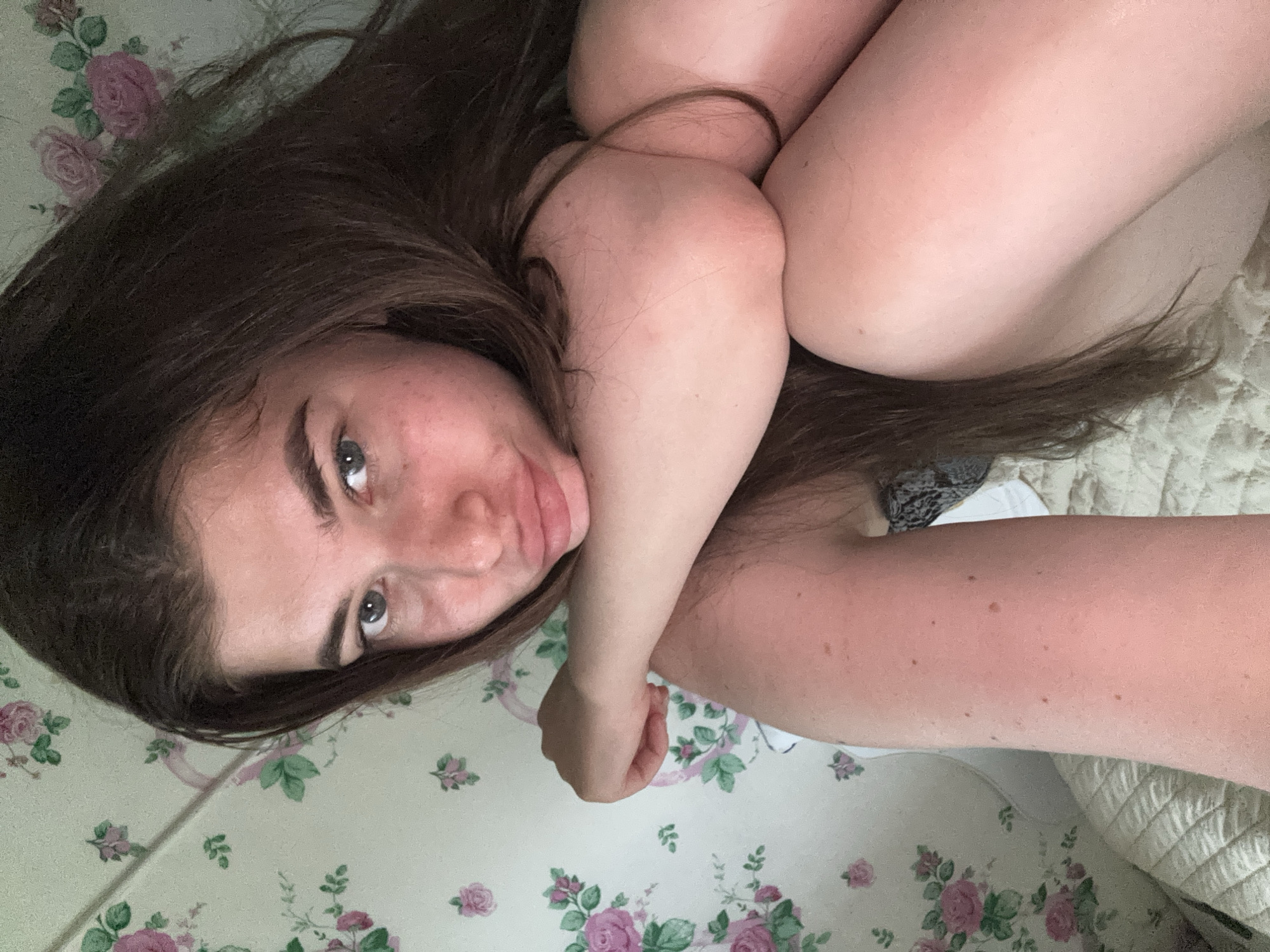 This is a horizontal, high-angle, close-up selfie of a young person lying down on a floral-patterned surface. The person has long, dark brown hair that falls around their face and body, partially obscuring their features. Their eyes are open and looking towards the camera, and their lips are slightly pursed. Their arms are wrapped around their legs, creating a somewhat curled-up pose. The background consists of a light-colored fabric with a repeating pattern of pink roses and green leaves. The overall lighting suggests it might be daytime, possibly indoors given the intimate setting. The person's expression appears relaxed or contemplative. There is no visible text in the image. The location context is Мещёры, Russia.