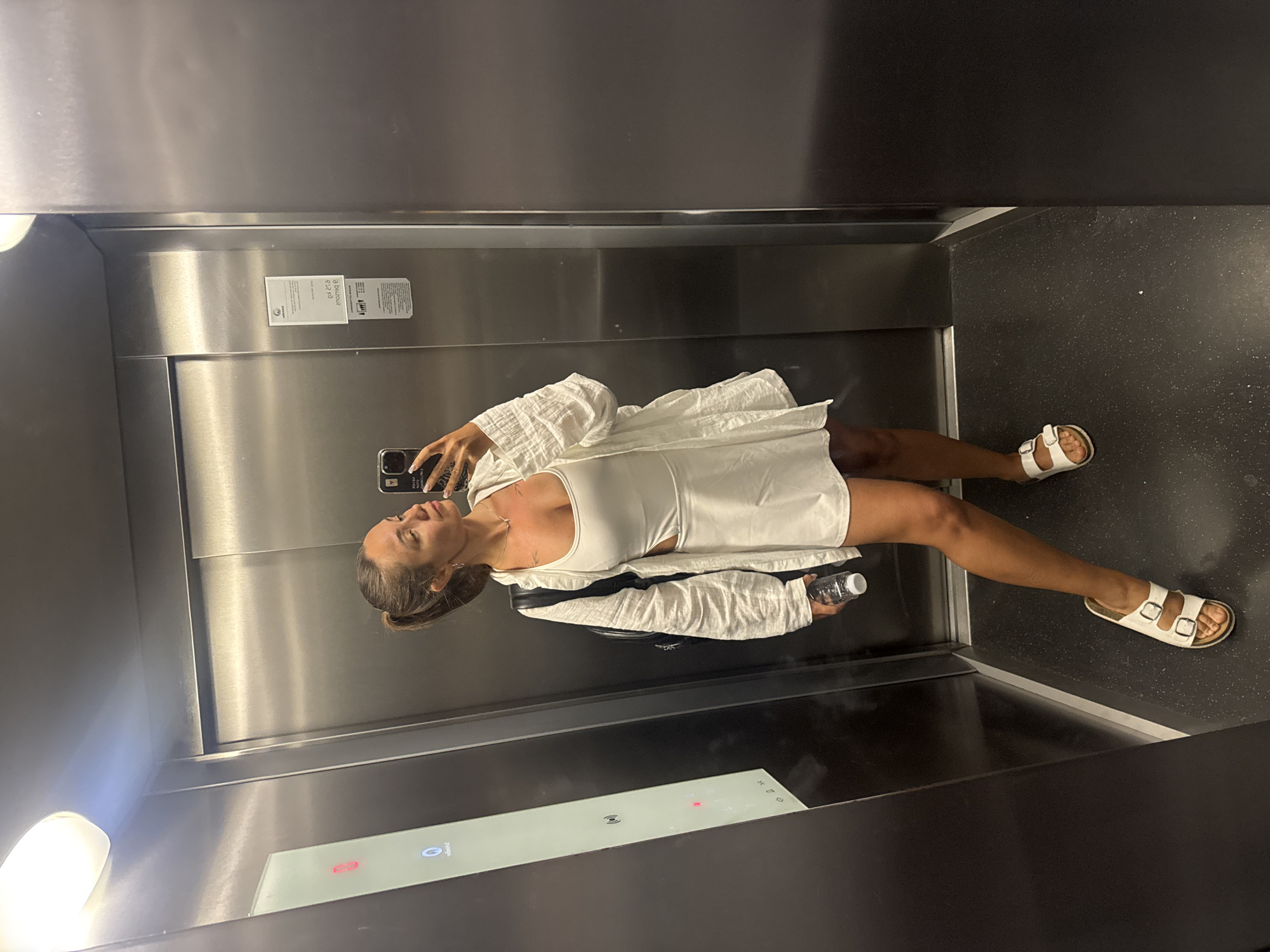 A woman is taking a mirror selfie in an elevator, likely in Budva, Montenegro, based on the provided context. She is wearing a white tank top, a white mini-skirt with a cutout on the side, and an unbuttoned white linen shirt over it. She has white Birkenstock sandals on her feet and her hair is tied in a ponytail. She is holding a clear water bottle in her right hand. The elevator has stainless steel walls and a dark floor. There are elevator controls visible on the left side of the image. The lighting in the elevator is bright, coming from an overhead light fixture.