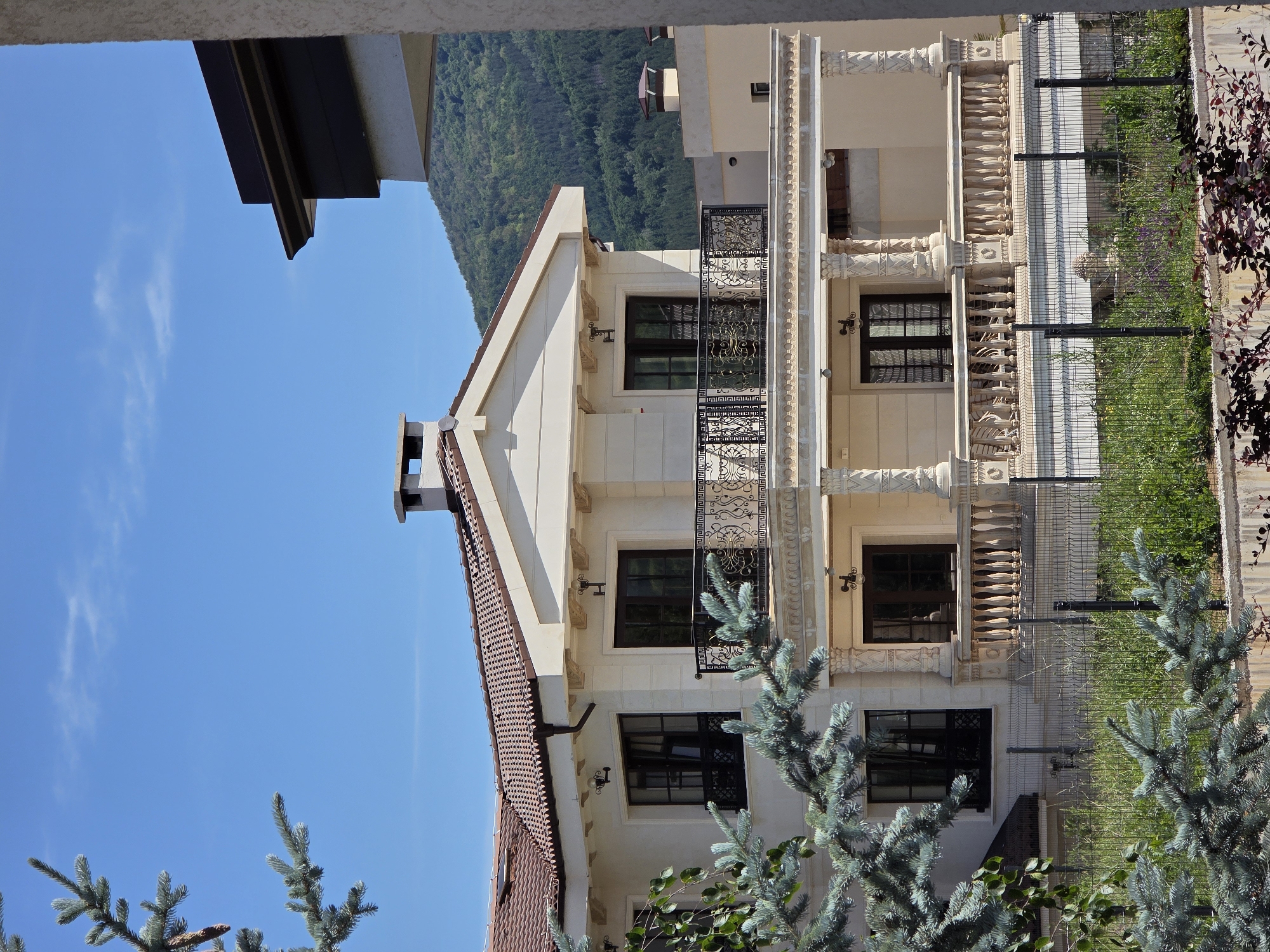 The image shows a large, modern, multi-story house with a tiled roof and light-colored walls. The house features several balconies with decorative iron railings and stone balustrades. Large windows and doors are visible. In the foreground, there are branches of a blue spruce tree and a wire fence. Behind the house, a forested mountain slopes upwards against a clear blue sky. The scene is situated in Kladnitsa, Bulgaria.