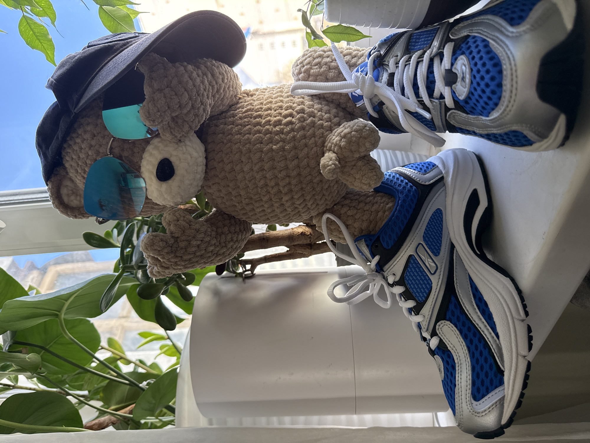 The image displays a large, light brown crocheted teddy bear or animal figure, positioned as if sitting or lounging by a window. The bear is accessorized with a dark baseball cap worn backward and cool blue mirrored sunglasses, reflecting parts of the bright sky and indoor elements. Its body shows a distinct crocheted texture.
The figure's feet are outfitted with blue, white, and silver athletic sneakers featuring white laces and mesh material. Small brand logos or text, possibly "Reebok" or similar, are faintly visible on the side and tongue of the shoes, along with small blue tags on the laces that are illegible.
The background reveals a sunny outdoor scene through a window, with a clear blue sky. Green foliage from houseplants partially frames the view. Beyond the plants and window, parts of light-colored, possibly stone buildings with some architectural detailing can be seen, suggesting an urban setting. The bear is placed on a white, flat surface, likely a window sill or shelf, with a white cylindrical object nearby. The bright natural light indicates it is daytime. The overall mood is playful and whimsical, portraying a charming, accessorized toy enjoying a bright day indoors in Chaikovsky, Russia.