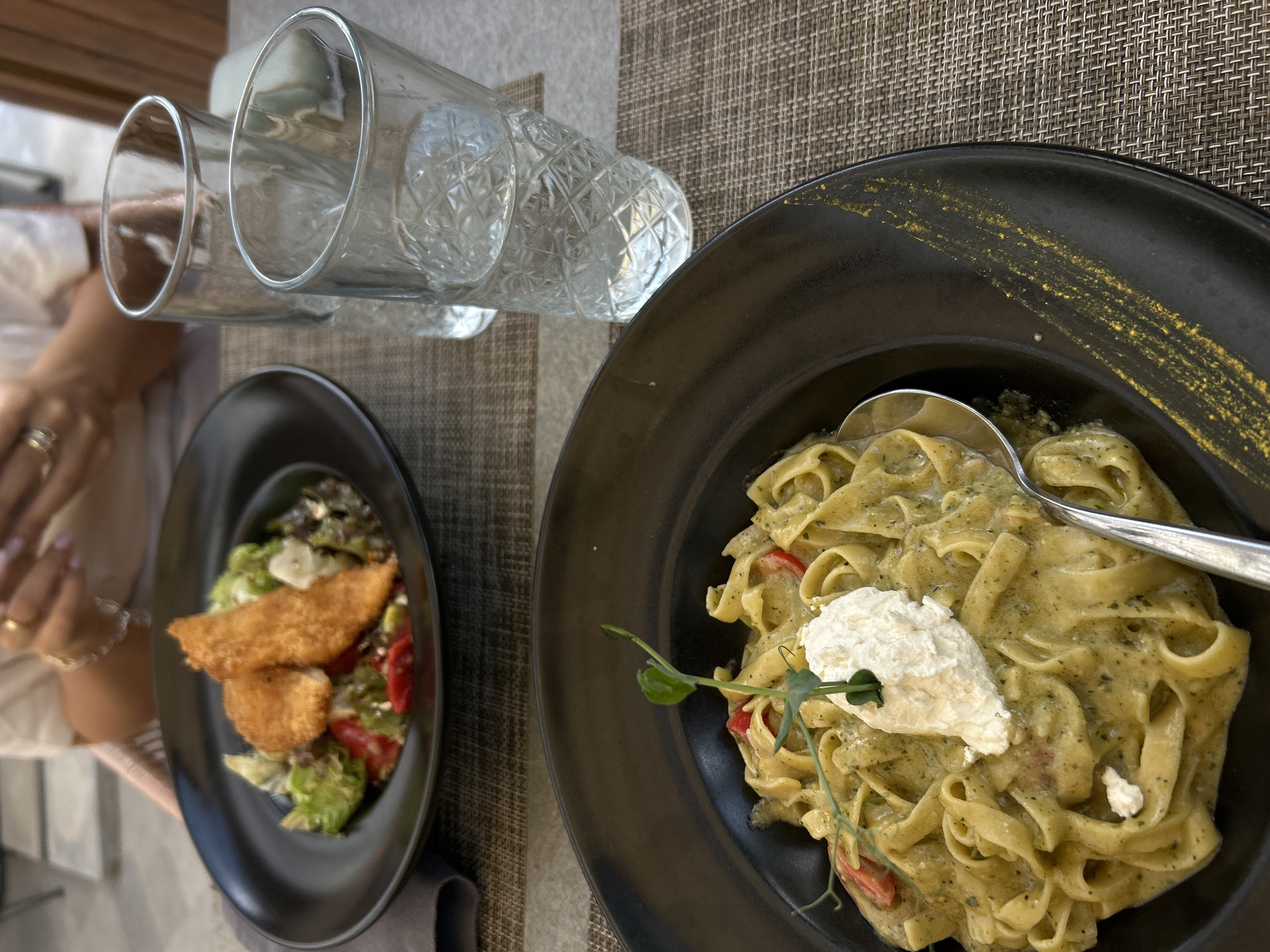 The image shows a table setting at a restaurant. Two clear glasses are placed near the top center of the frame. A black plate with a salad and fried breaded items is positioned on the left, partially obscured by a person's arm wearing rings. On the right, a larger black plate contains pasta with a green pesto-like sauce and a dollop of white cream or cheese, with a spoon resting on top. A small garnish of green leaves is visible on the pasta. The table surface is covered with a textured, neutral-toned material. The location is Sofia, Bulgaria.