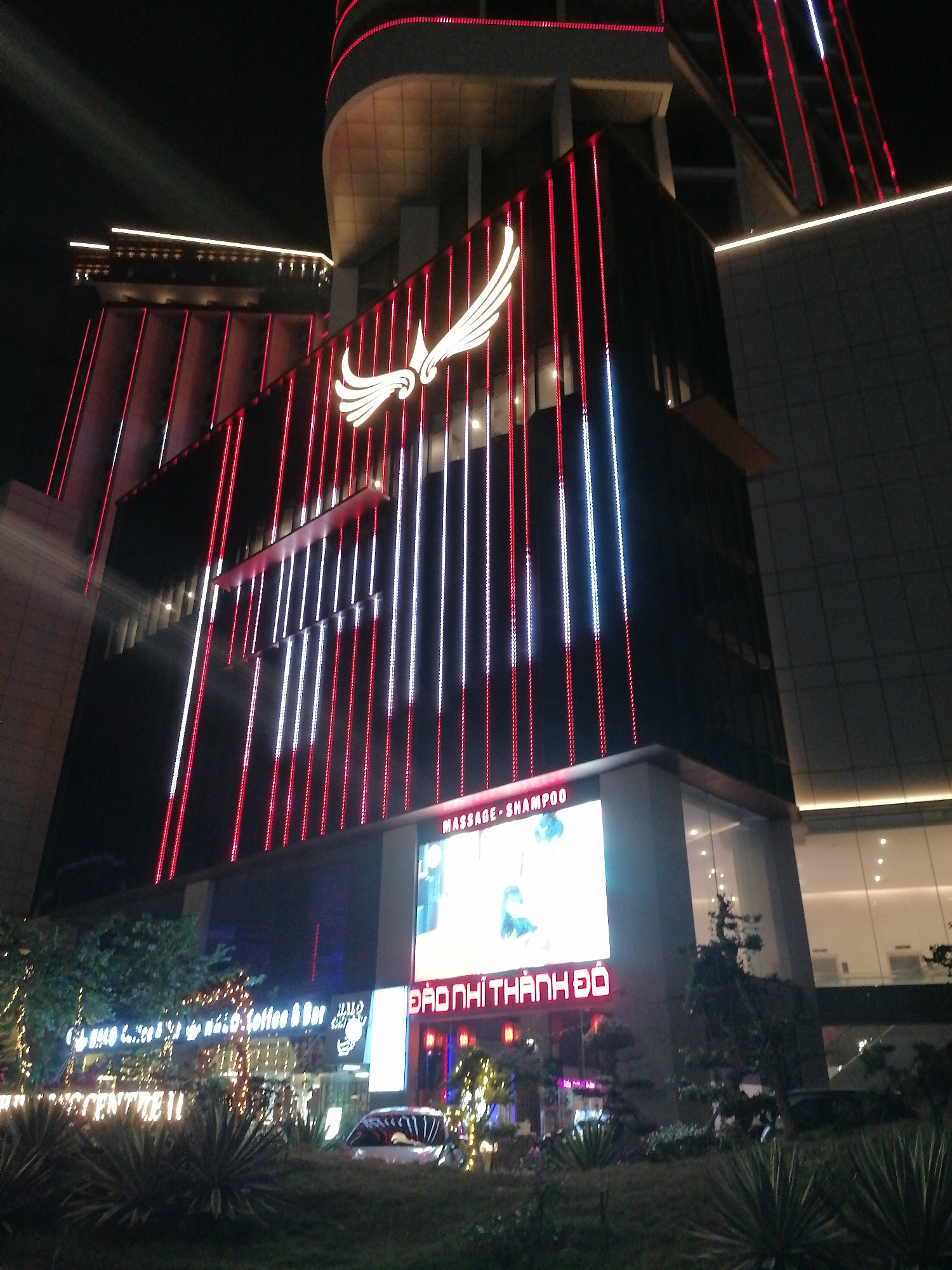 This is a nighttime photograph of a modern, multi-story building in Bai Chay Ward, Vietnam. The building is extensively illuminated with vertical strips of red and white LED lights, creating a dynamic visual effect on its facade. A large, white, stylized winged logo, possibly representing a phoenix or eagle, is prominently displayed near the top of the illuminated section.
At the lower levels of the building, a large digital display screen is visible, showing the English text "MASSAGE - SHAMPOO" at the top and the Vietnamese text "ĐẠO NHỊ THÀNH ĐÔ" in red below. To the left, another commercial establishment, adorned with string lights, shows partial text including "Coffee & Bar" and "CENTRE", suggesting a cafe or bar.
The foreground features lush, manicured greenery, including various bushes and small trees, with some ground lighting. A white car is partially visible, parked amidst the foliage on the right side of the frame. The sky is dark, indicating that the photo was taken at night. No specific human activity or event is clearly depicted. The scene captures the vibrant, illuminated urban landscape of the area.