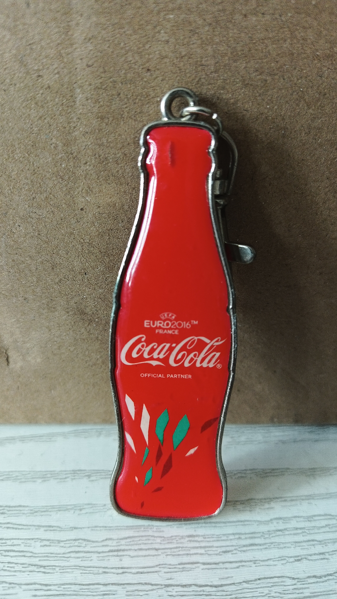 A close-up shot depicts a Coca-Cola keychain against a neutral background. The keychain is shaped like a classic Coca-Cola bottle, filled with red enamel. The Coca-Cola logo is featured prominently. Above it are the words "UEFA EURO2016 FRANCE". Underneath, the keychain states "OFFICIAL PARTNER". A scattering of stylized green and white designs are present at the bottom. A metal ring is attached to the top. The keychain rests on a light-colored surface with linear textures.