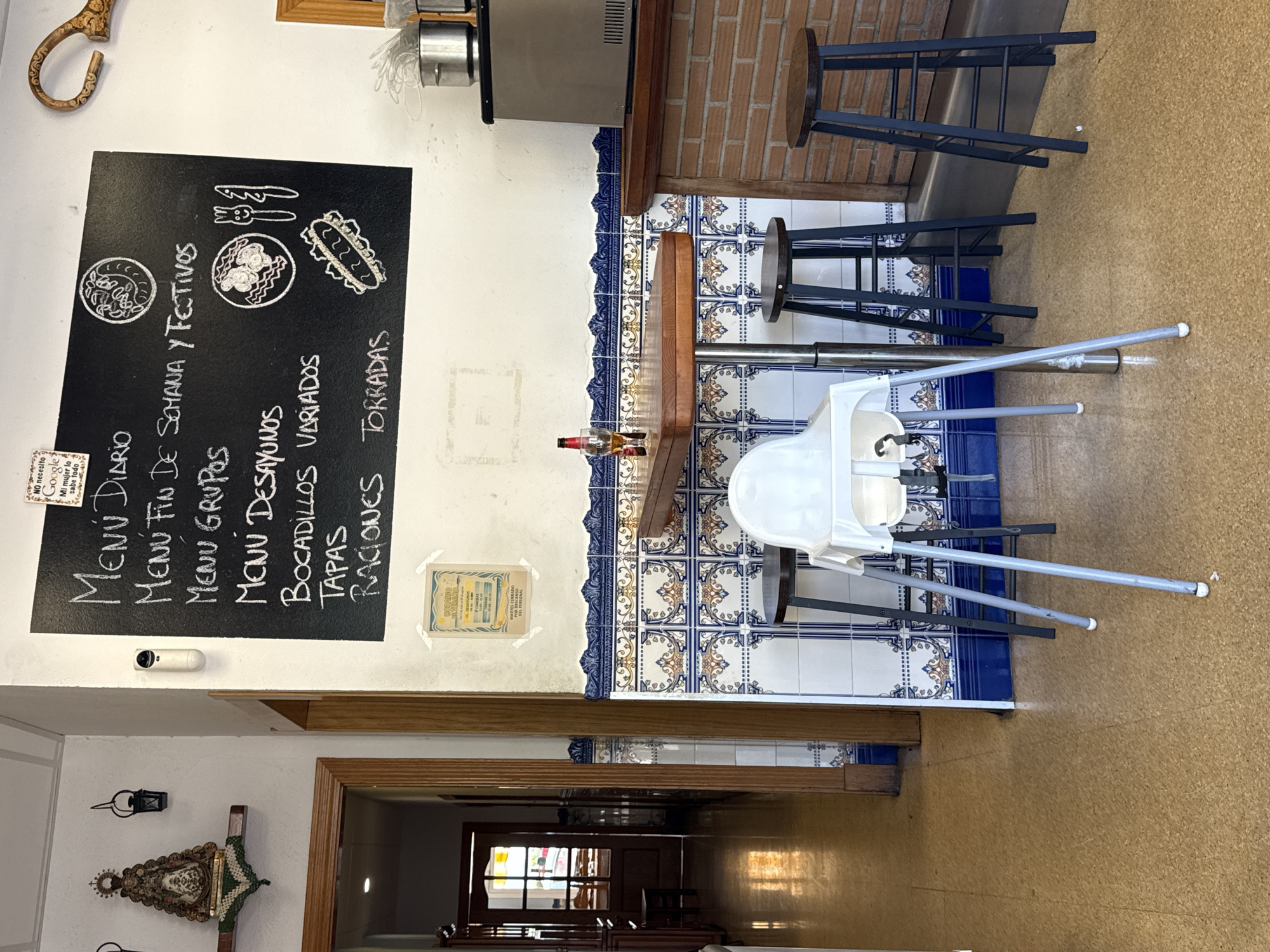 The image shows the interior of a casual dining establishment, likely a tapas bar or restaurant, in Blanes, Spain. A prominent blackboard menu is displayed on the wall, listing various menu options in Spanish: "Menú Diario," "Menú Fin De Semana Y Festivos," "Menú Grupos," "Menú Desayunos," "Bocadillos Variados," "Tapas," and "Raciones Torradas." Decorative chalk drawings of food items and utensils are also present on the blackboard.
In the foreground, a high chair for a child is positioned next to a small, round wooden table with a metal pedestal base. On the table, a glass of amber liquid and a bottle of beer are visible. Two tall stools with dark wooden seats and black metal frames surround the table. A third similar stool is located to the right, closer to a brick counter.
The walls are decorated with a distinctive blue and white tile pattern along the lower section, and plain white above. A framed sign reading "Horario de Verano" (Summer Schedule) is attached to the wall above the tiles. In the background, a doorway leads to another part of the establishment, with warm sunlight filtering through. To the left, on a shelf, a religious figurine of the Virgin Mary is displayed with a small, dark lantern hanging beside it. The floor is a light brown, speckled material. A metal appliance, possibly a refrigerator or a coffee machine, is situated on the brick counter. The overall ambiance suggests a relaxed and family-friendly dining environment.