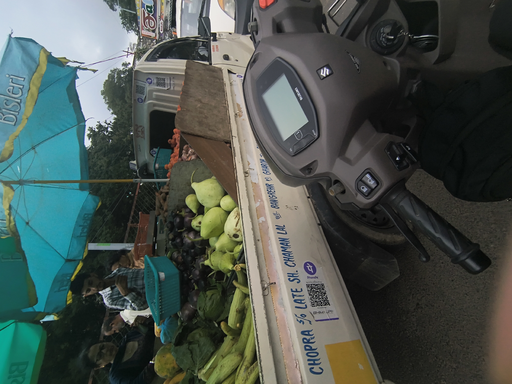 A street-side vegetable market is depicted, likely in Dharamshala, India, given the presence of a "CHOPRA S/O LATE SH. CHAMAN LAL" inscription on the side of a truck, a common naming convention in North India. In the foreground, a dark-colored scooter is parked, with its handlebars and instrument panel visible. Behind the scooter, a white pickup truck is loaded with a variety of fresh produce, including green gourds, eggplants, and leafy vegetables. A person in a plaid shirt is interacting with the vegetables, presumably a vendor. Two women, one wearing glasses, are also present near the produce stall. Blue and yellow umbrellas provide shade over the market. The scene suggests an active, open-air marketplace.
