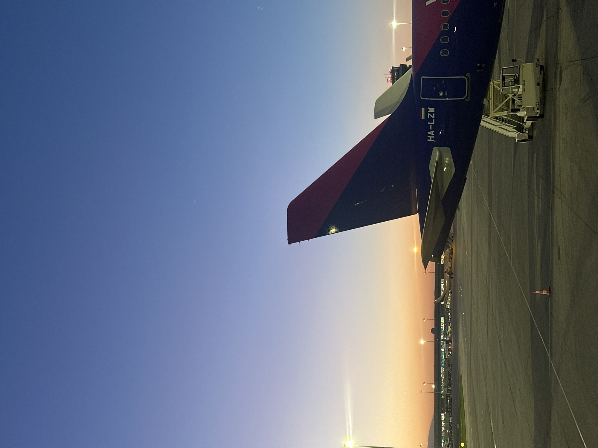 The image shows a Wizz Air airplane, registration number HA-LZW, parked at an airport. The tail of the plane is visible, painted with red and black. A mobile boarding stair is attached to the plane. The scene appears to be at Sofia Airport in Sofia, Bulgaria. In the background is an airport apron with other airplanes parked, and streetlights are visible. The sky is a gradient of blue to orange, suggesting dusk or dawn.