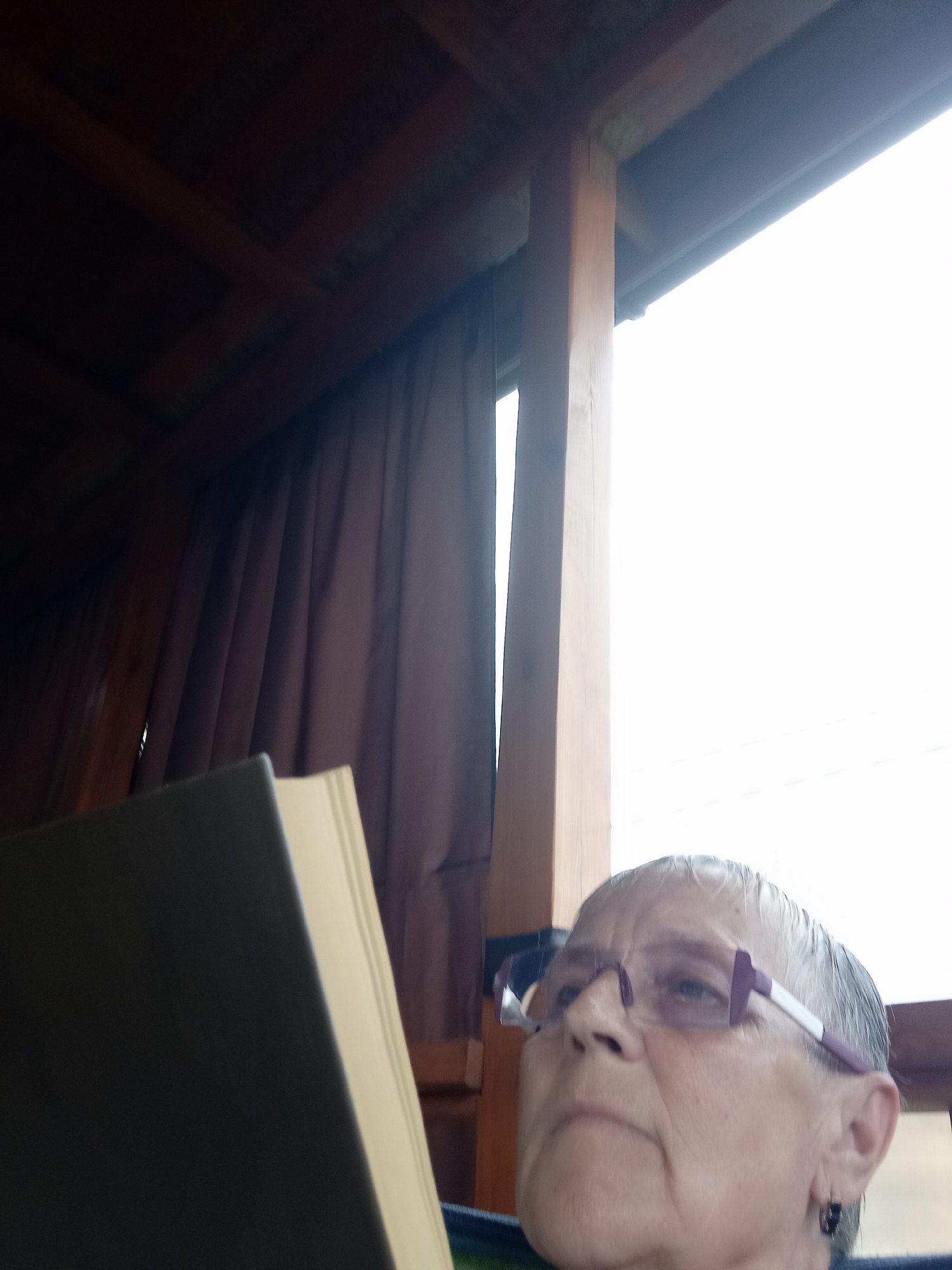 The image shows a woman wearing glasses and reading a book. She is sitting outdoors, under a wooden roofed structure, possibly a porch or gazebo. The background features brown curtains and a bright, blown-out sky. The woman is looking down at her book, with her face partially illuminated by natural light. The scene appears to be from a residential setting, and there are no discernible landmarks to pinpoint a specific location within Degtyarsk, Russia.