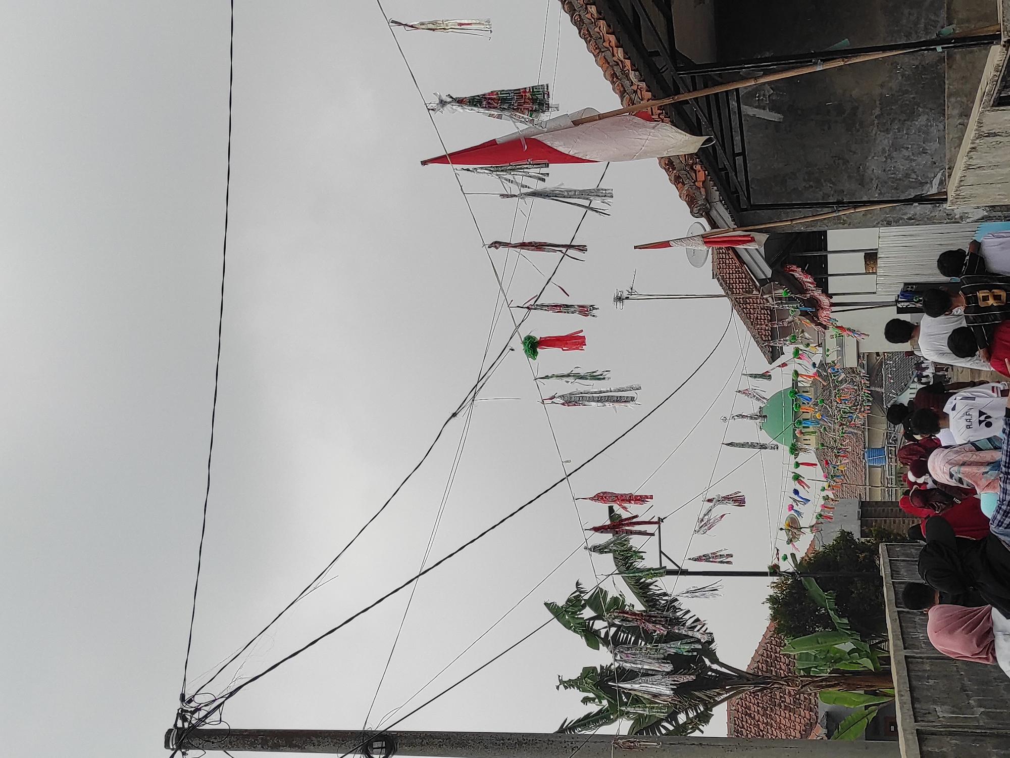 The image depicts a street scene in Sumedang, Indonesia, likely during a festive occasion. The sky is overcast. Overhead, a tangle of electrical wires is strung across the scene, adorned with numerous colorful banners and decorations. These decorations appear to be in the form of small flags, streamers, and other festive ornaments, some resembling tassels. A prominent Indonesian flag, red and white, is visible flying from a pole on the right side of the image, attached to a wire.
In the mid-ground, a green domed building, possibly a mosque or a significant public structure, is visible with more decorations strung along its perimeter and overhead. Trees with broad leaves, likely banana trees, are present on the left side.
In the foreground, a group of people, including women wearing hijabs and men and children in casual attire, are walking away from the camera, heading down the street. Some of the children are wearing athletic clothing, one with the number '8' visible on his back. The street itself appears to be paved, with a concrete wall or barrier on the right side. Buildings with red tiled roofs are visible throughout the scene, suggesting a residential or mixed-use area. The overall impression is one of community celebration and public decoration in an Indonesian urban or semi-urban setting.