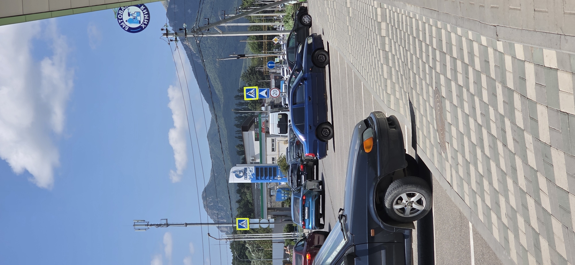 A blue sedan is parked in a lot in front of a gas station. There are several other cars in the lot and on the road. In the background, there are mountains and a cell tower. A sign for "Gazprom" is visible above the gas station. The image is taken on a sunny day with a blue sky and some clouds.