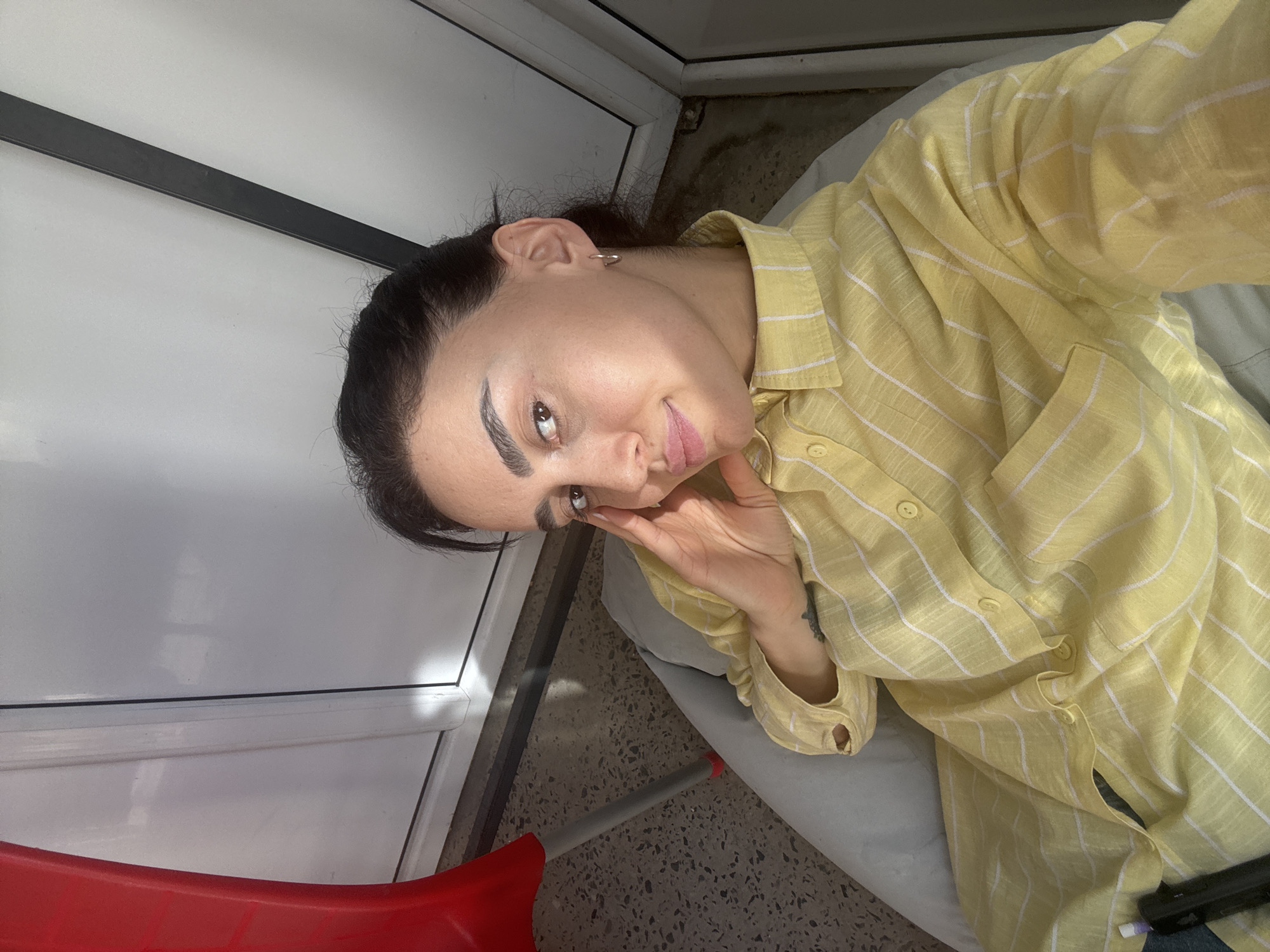 A woman is shown indoors in a selfie-style shot, wearing a yellow and white striped button-down shirt. She has dark hair pulled back, fair skin, and is wearing small hoop earrings. Her left hand is touching her face near her eye. Behind her is a white door frame with a black accent stripe, and a speckled floor. A red object, possibly part of a chair or cleaning tool, is visible in the bottom left corner. The location is likely indoors in Tbilisi, Georgia, as requested, although specific architectural features or indicators are absent from the image to confirm the city definitively.