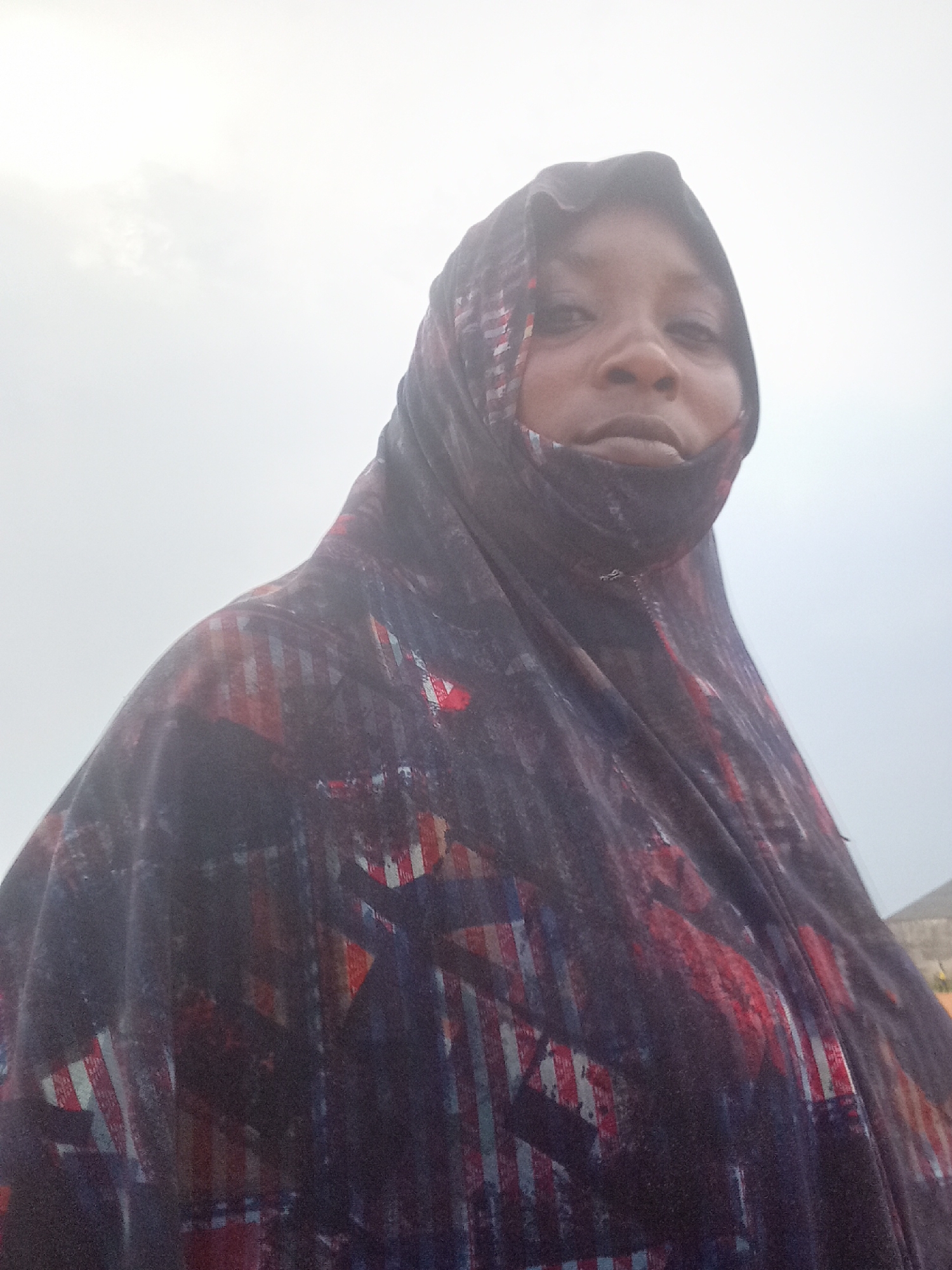A medium close-up shot depicts a woman wearing a dark, patterned hijab. The hijab features red, white, and blue linear patterns. The woman's face is visible, with neutral expression. The background is a bright, overcast sky. The location is in the city of Akwanga, Nigeria.