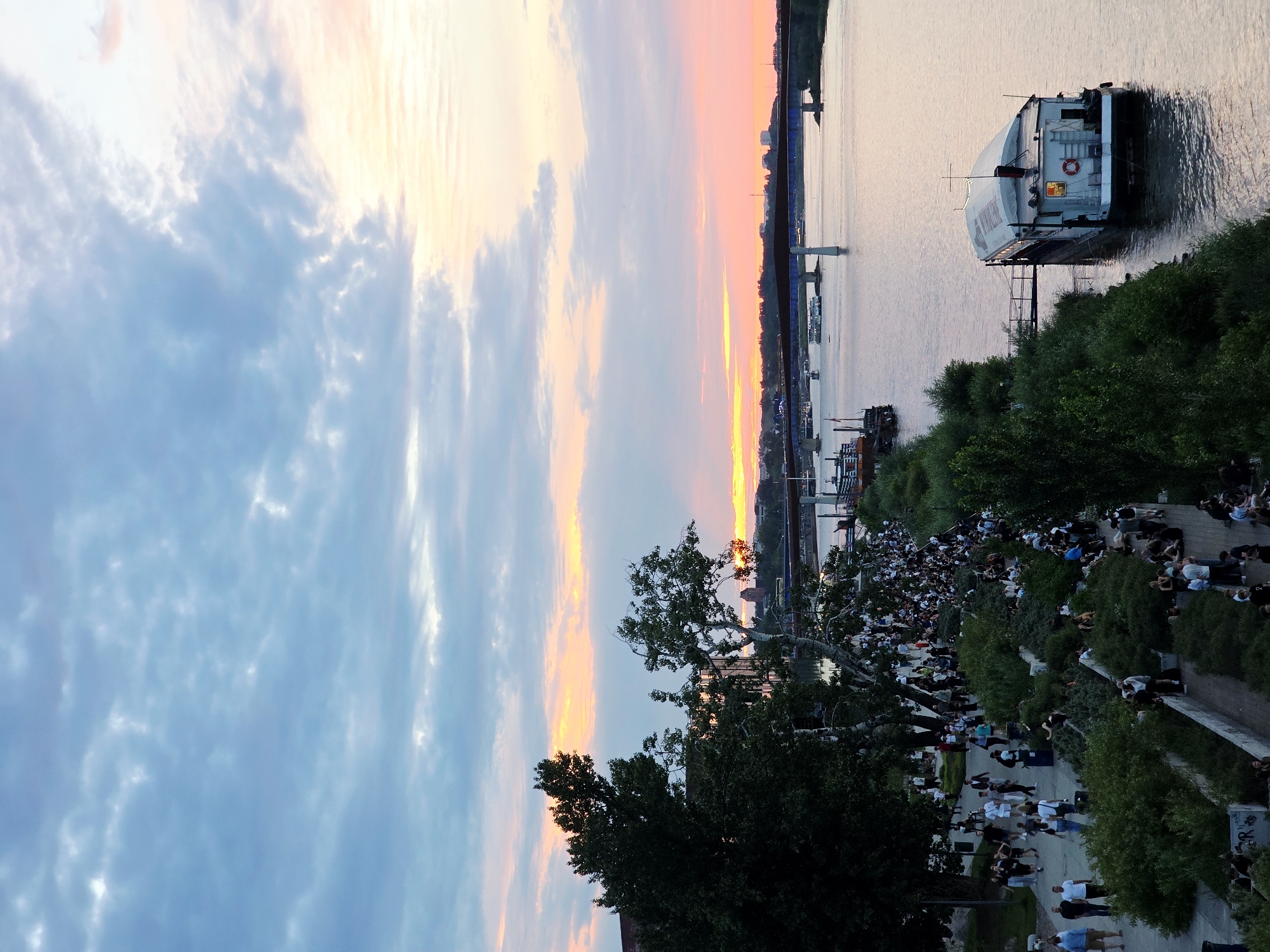 The image captures a scene along the Vistula River in Warsaw, Poland, during a sunset. A pedestrian promenade is visible on the left, lined with trees and bushes, where a large number of people are gathered, some walking and others sitting on the grass and paved areas. Further into the distance, a bridge spans the river, with several boats navigating the water. A houseboat is moored to the embankment in the foreground, its windows illuminated. The sky is filled with clouds, displaying hues of blue, pink, and orange as the sun sets.