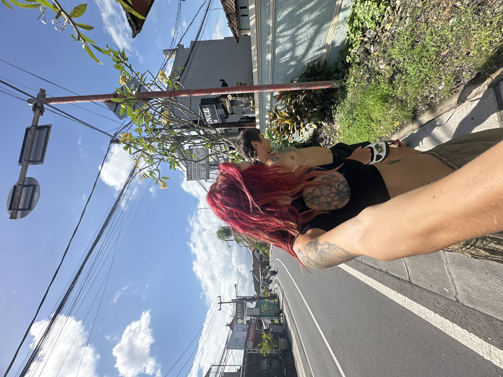 A street-level, upward-angled shot captures a man and a woman walking alongside a road, likely in North Kuta, Indonesia. The woman has long, red hair and a tattoo on her back; she is holding a retractable dog leash. The man beside her is shirtless. The environment includes tropical foliage, buildings, utility poles with numerous wires against a blue sky with scattered clouds, and street signs written in Bahasa Indonesian. The road shows signs of pedestrian activity and vehicular traffic.
