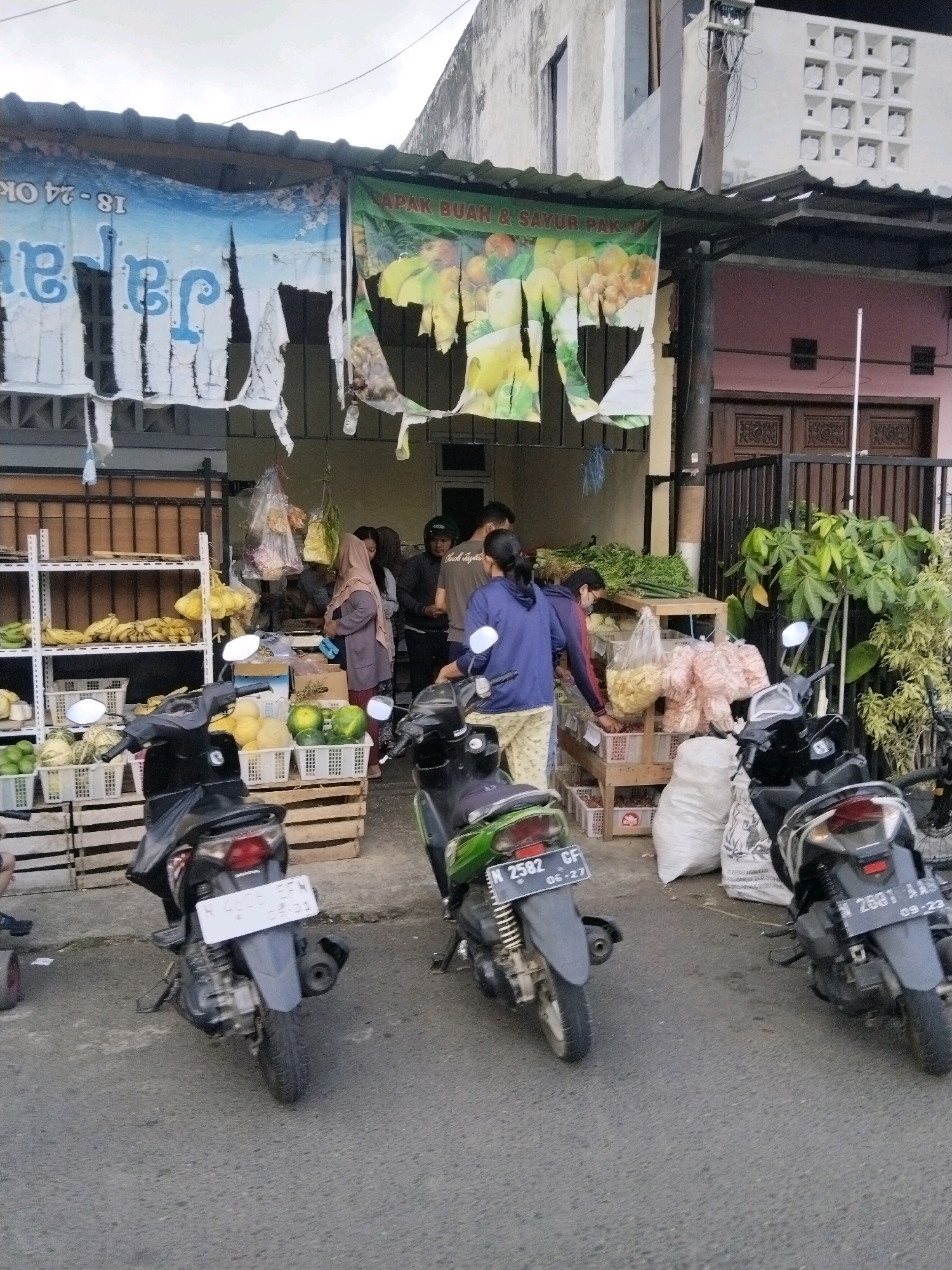 The image depicts a street-side fruit and vegetable stall in Banjararum, Indonesia. A covered stall displays produce on shelves and in crates. Bananas, melons, and leafy green vegetables are visible. Several people are interacting within or near the stall, some appearing to be customers and others vendors. Three scooters are parked in front of the stall on the paved road. Banners with Indonesian text are hung above the stall. The environment suggests an outdoor marketplace setting with buildings in the background.