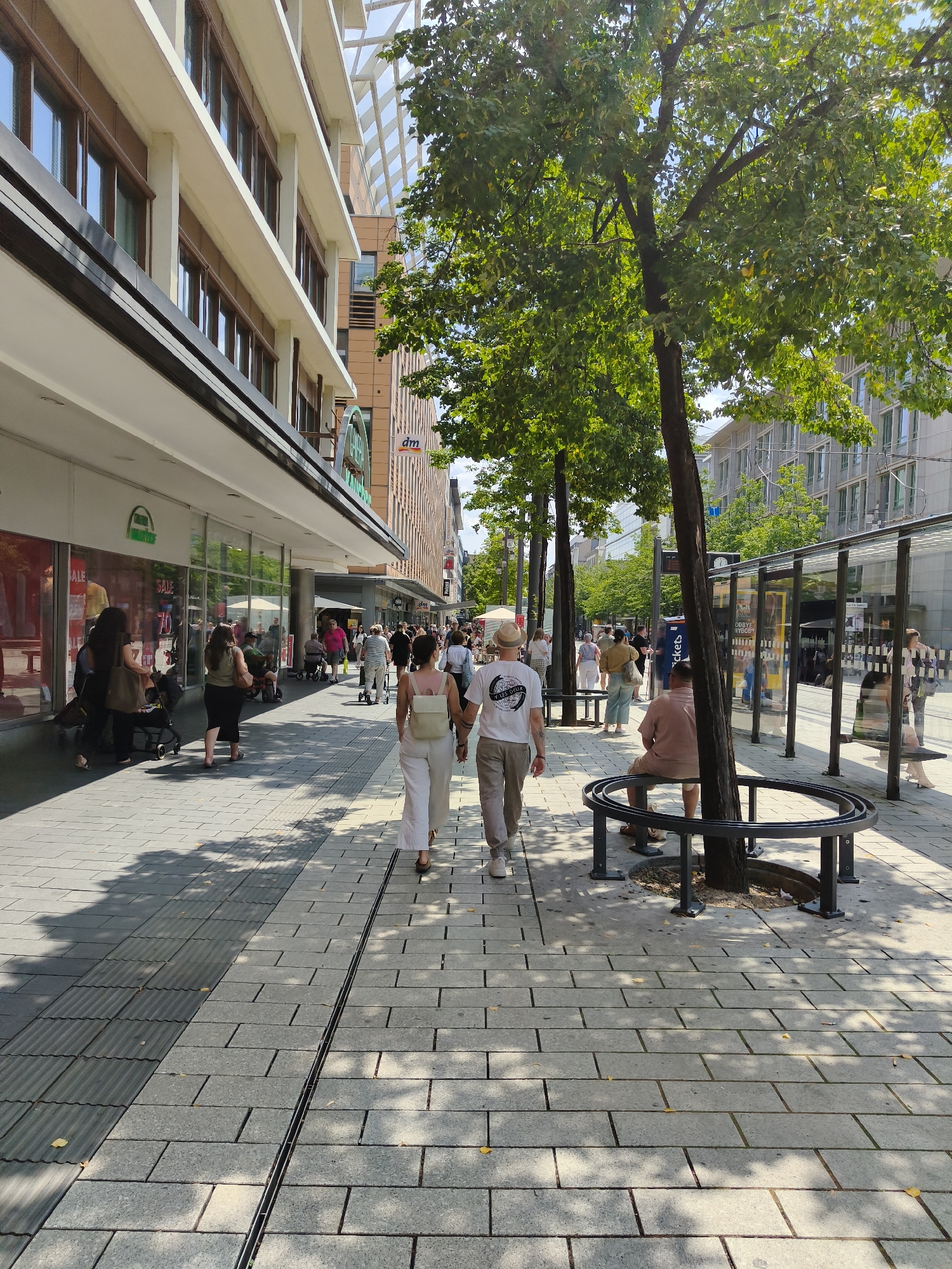 The image depicts a street scene in Mannheim, Germany, specifically on Planken, a pedestrian shopping street. The foreground shows a paved sidewalk with a pattern of grey rectangular stones and a drainage channel. Sunlight casts dappled shadows from trees onto the ground. Two individuals are walking away from the camera on the sidewalk. The woman in front wears white linen trousers and a light-colored top with a backpack. The man behind her wears beige trousers and a white t-shirt.
To the left of the sidewalk is a row of shops with large glass windows. Signs visible on the shopfronts include "SALE" and "70". A green logo is also visible. Further down the street, more people are walking and shopping.
To the right of the sidewalk, there is a bus stop with a glass enclosure and seating. A tree trunk is prominent in the middle ground, with its branches and green leaves extending into the upper part of the frame. Behind the bus stop and trees, modern buildings with multiple windows are visible. In the background, other buildings and more trees line the street. The overall scene suggests a busy, sunny day in a commercial district.