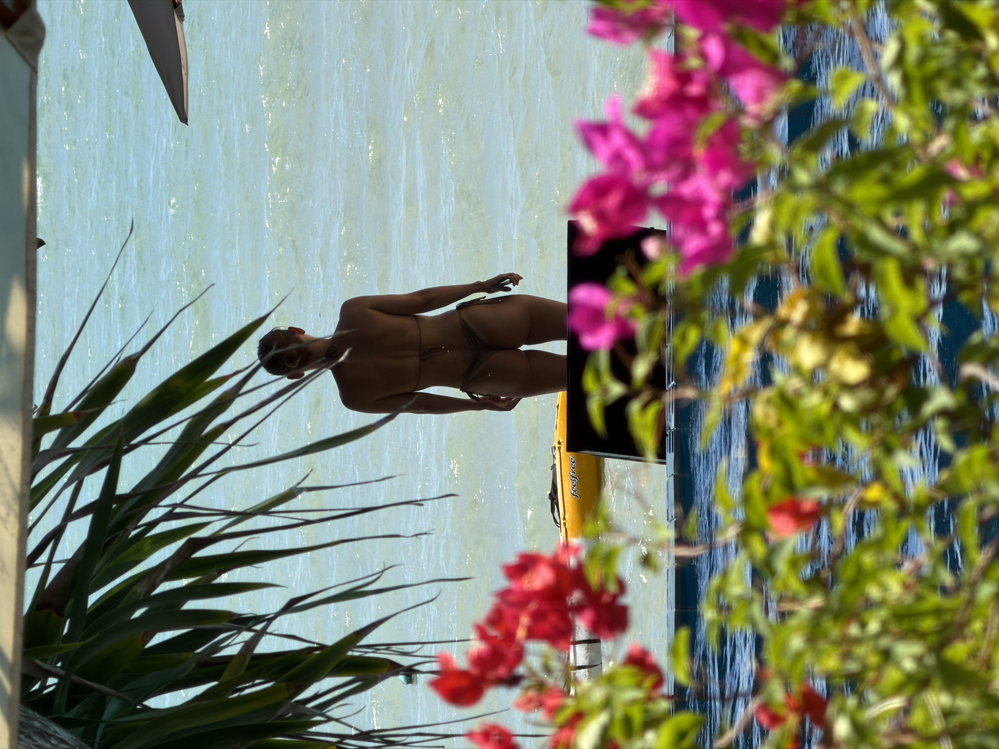A woman in a bikini stands on a beach in Ko Samui District, Thailand, facing the ocean. In the foreground, bougainvillea flowers and palm leaves partially obscure the view. A yellow kayak is visible in the water behind the woman. The sun is shining, and the ocean appears calm with gentle waves. The overall scene suggests a relaxed tropical vacation atmosphere.