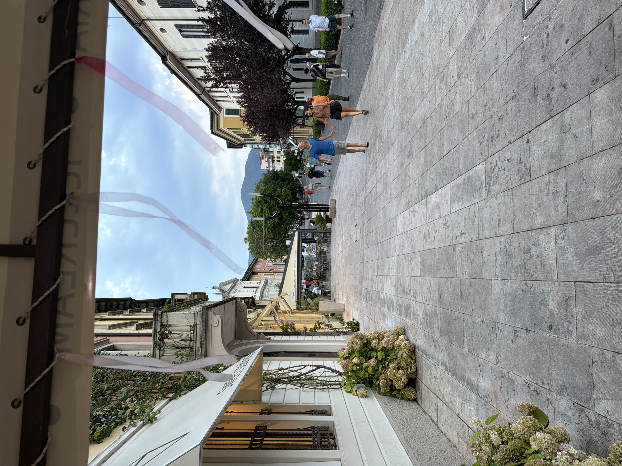 The image depicts a street scene in the city of Salò, Italy, likely on a pedestrian walkway. In the foreground, a light-colored tiled pavement stretches into the distance. To the left, a building with a white facade and awnings is visible, with a large cluster of light-colored hydrangeas at its base. Further down the street, buildings with various colored facades line both sides. On the right, several trees with dark foliage are present.
Several people are walking along the street. In the mid-ground, a couple is holding hands and walking towards the viewer. The man is wearing a blue t-shirt and khaki shorts, while the woman is wearing a brown top and dark shorts. Other individuals are scattered further along the walkway, some walking, others standing.
Above the scene, the sky is overcast with some clouds. White and pink ribbons are hanging from above, possibly as decoration. The overall impression is that of a casual daytime stroll in a European town.