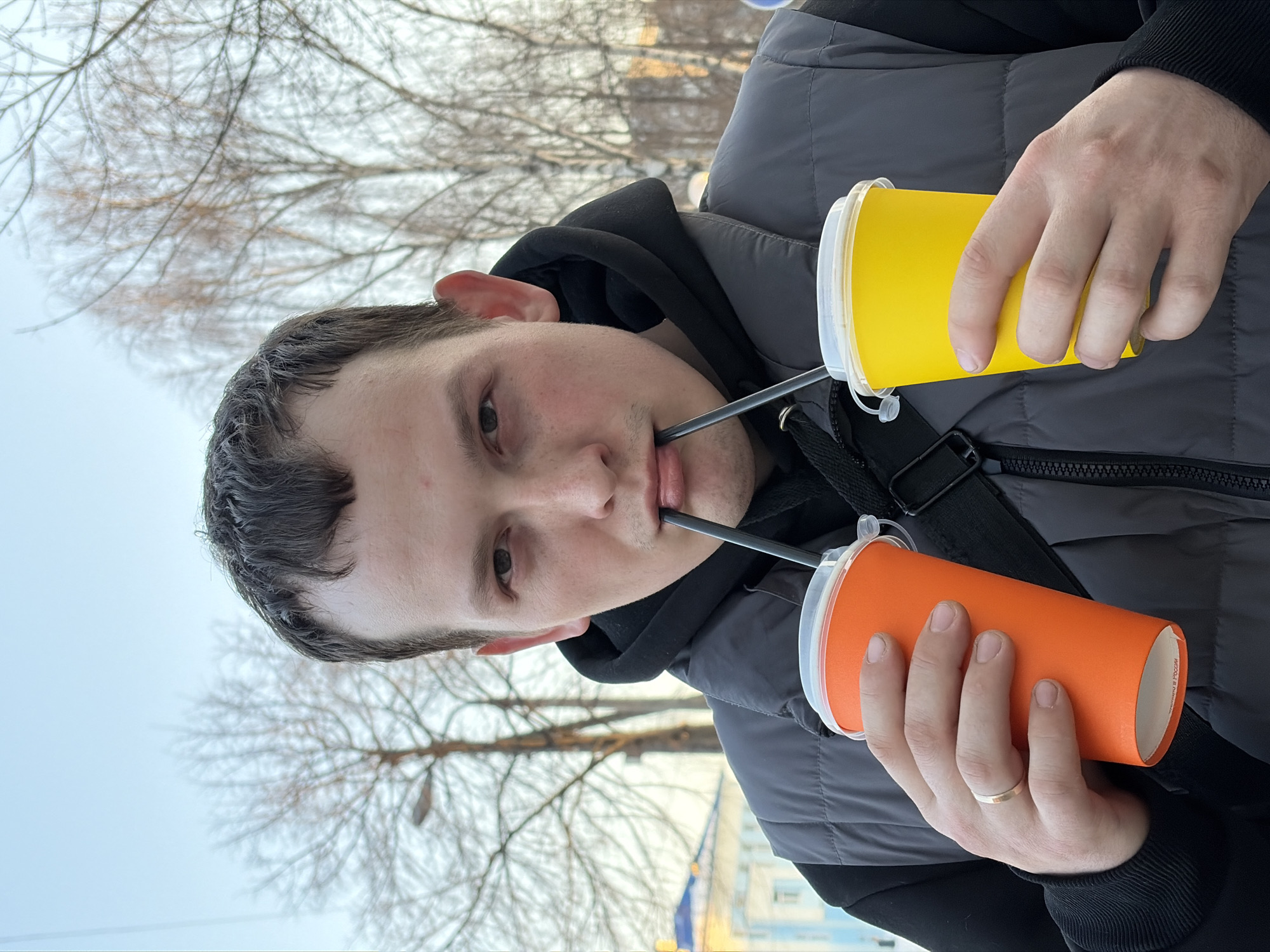A male individual, appearing in his 20s or 30s, with short dark hair and a light complexion, is shown from the chest up, looking directly at the camera. He is wearing a dark gray sleeveless puffer vest over a black hooded sweatshirt. A gold band ring is visible on the ring finger of his left hand. He is simultaneously holding an orange paper cup with a clear plastic lid and black straw in his left hand, and a yellow paper cup with a clear plastic lid and black straw in his right hand. Both black straws are inserted into his mouth. The scene is outdoors under a light blue sky, with several bare deciduous trees in the background, indicative of late autumn, winter, or early spring. A street light or utility pole is visible among the trees. To the far left, a light blue multi-story building with a dark blue pitched roof and white architectural elements is partially visible. The location is an urban setting in Chaikovsky, Russia; however, a precise street or address cannot be determined from the visual information provided.
