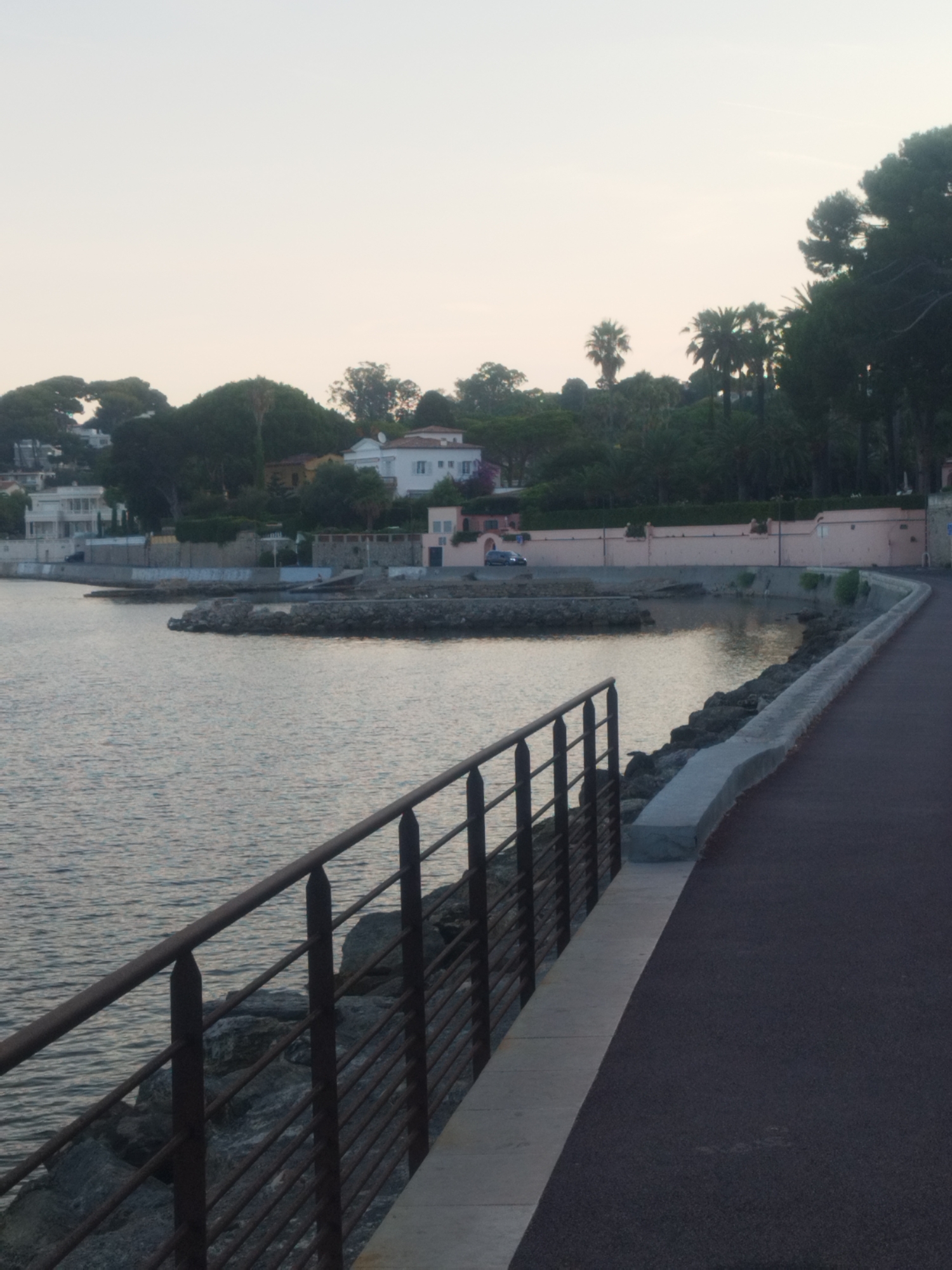 The image depicts a coastal scene in Antibes, France, likely during early morning or late evening due to the soft, diffused light. In the foreground, a dark asphalt path curves along the right side of the frame, separated from the water by a low concrete barrier and a dark metal railing. The railing, composed of vertical posts and horizontal bars, borders the pathway and overlooks a rocky shoreline. The water is calm, reflecting the muted sky.
Beyond the shoreline, a man-made harbor or cove is visible, with several rocky outcrops and structures within it. In the background, a series of buildings and vegetation are present. The buildings appear to be residential, with varying architectural styles, including white villas with tiled roofs and a longer pink structure. Lush green trees and palm trees dot the landscape, suggesting a Mediterranean climate. A dark-colored car is parked near one of the buildings. The overall atmosphere is tranquil and serene, characteristic of a seaside promenade.