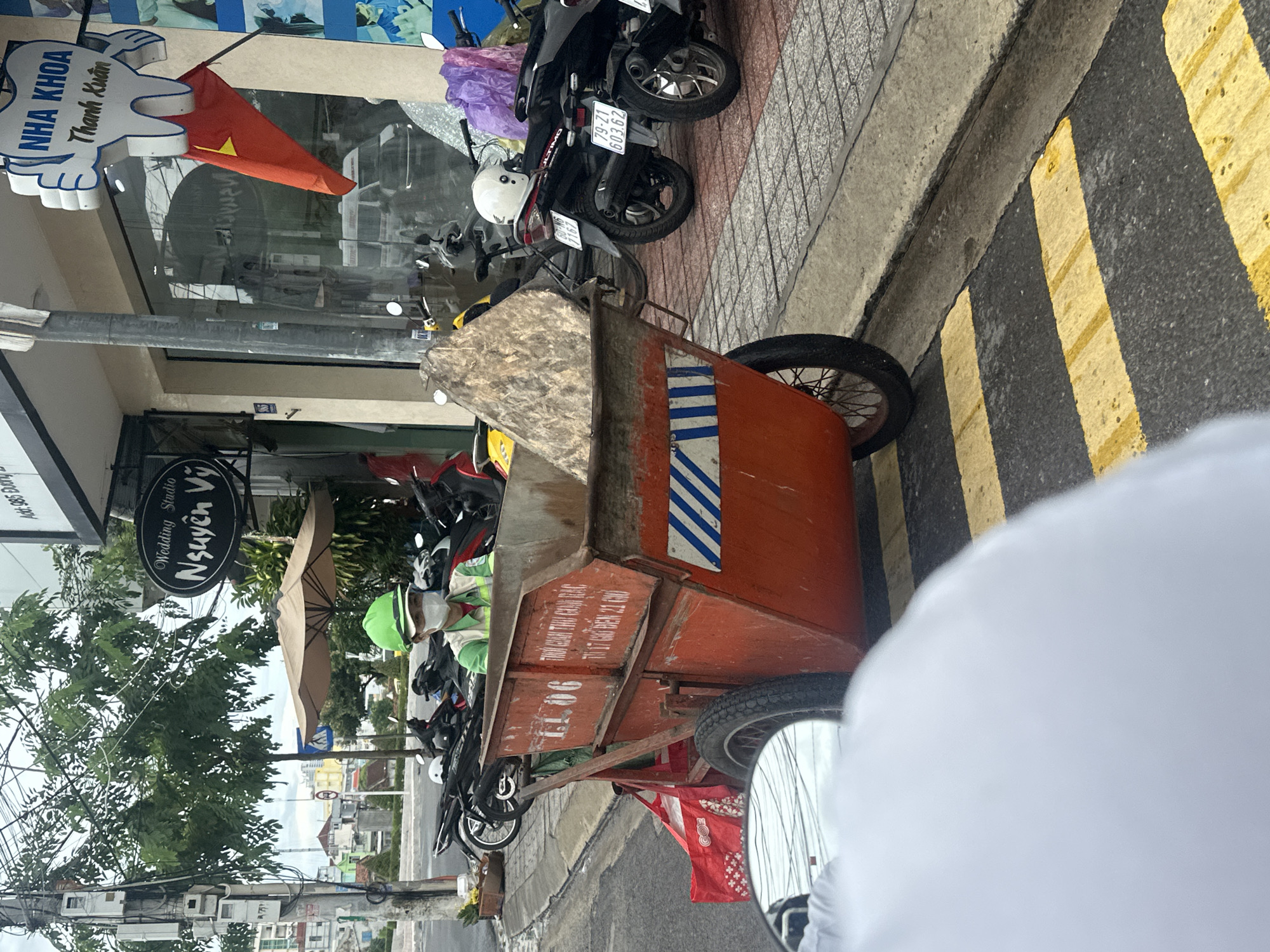 The image shows a street scene in Nha Trang Ward, Vietnam, focusing on a three-wheeled trash cart on the side of the road, parked next to a curb with yellow and black stripes. Several motorbikes are parked in front of a building, one of which is in front of the 'NHA KHOA Thanh Xuan' dental clinic. The “Wedding Studio Nguyen Vy” signage is also visible. An umbrella shades an area further down the street where people in green shirts and helmets are seen near the parked motorcycles. The trash cart is orange and has blue and white stripes, and the label "11.06" is printed on the side. The trash cart also includes printed text "THUOM CEN THE CASA RAC" along with smaller text that is less legible. A Vietnamese flag is hung near the dental clinic. The license plate on one of the motorbikes shows "79-Z1 603.62" indicating the vehicle is from Khánh Hòa Province where Nha Trang is located.