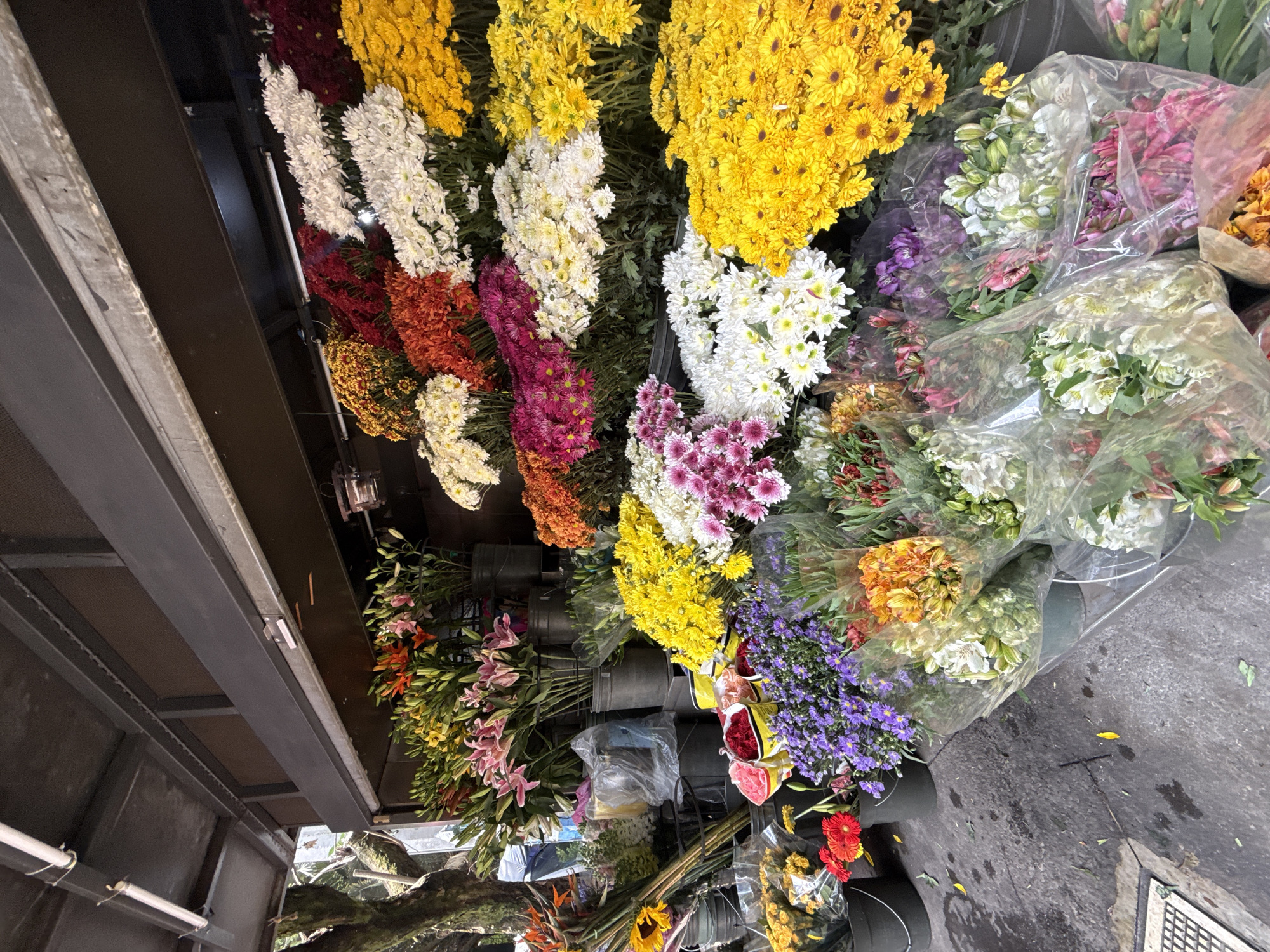 The image shows a flower stand at a market in Mexico City, Mexico. It features a variety of colorful flowers, including yellow and white chrysanthemums, purple asters, lilies, alstroemeria, and other mixed bouquets. Some of the flowers are in buckets and some are wrapped in clear plastic. The stand is under a dark metal overhang, and the floor is a concrete surface with some debris.